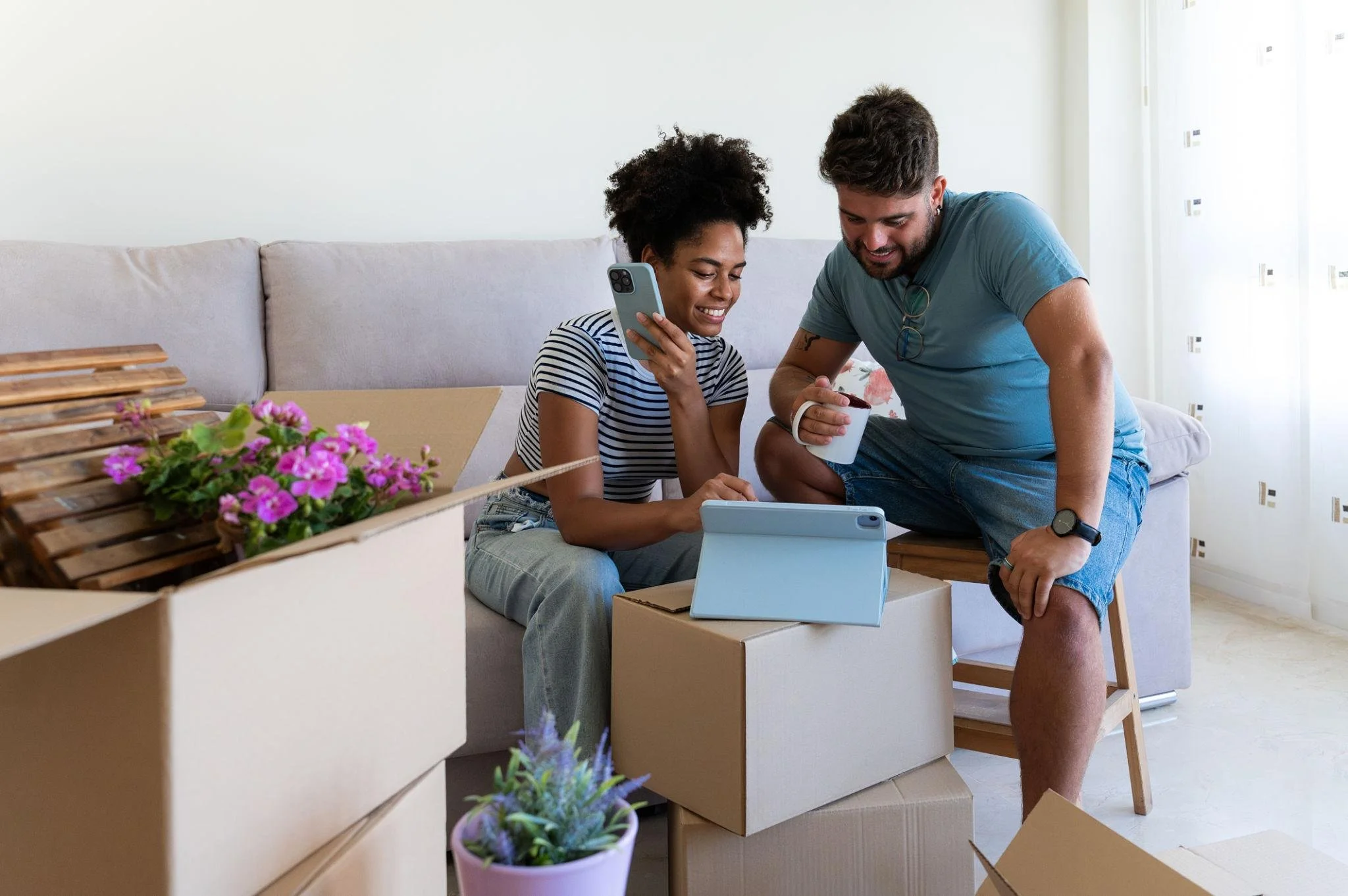 A smiling woman and man sitting on a sofa amid unpacked boxes, looking at a tablet together in a bright living room.