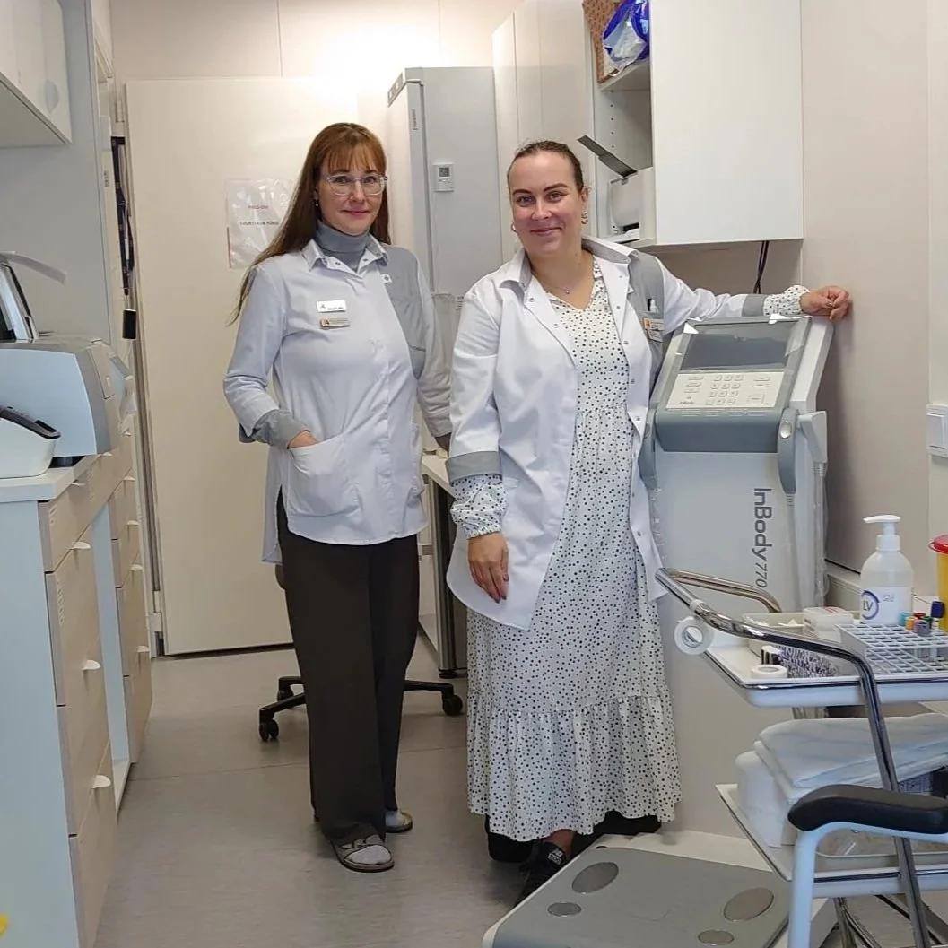 Kaksi naista työskentelee laboratoriossa, toinen kädellään laitteessa ja toinen seisoo vierellä. Molemmilla on laboratorioliivit ja he ovat hymyileviä.