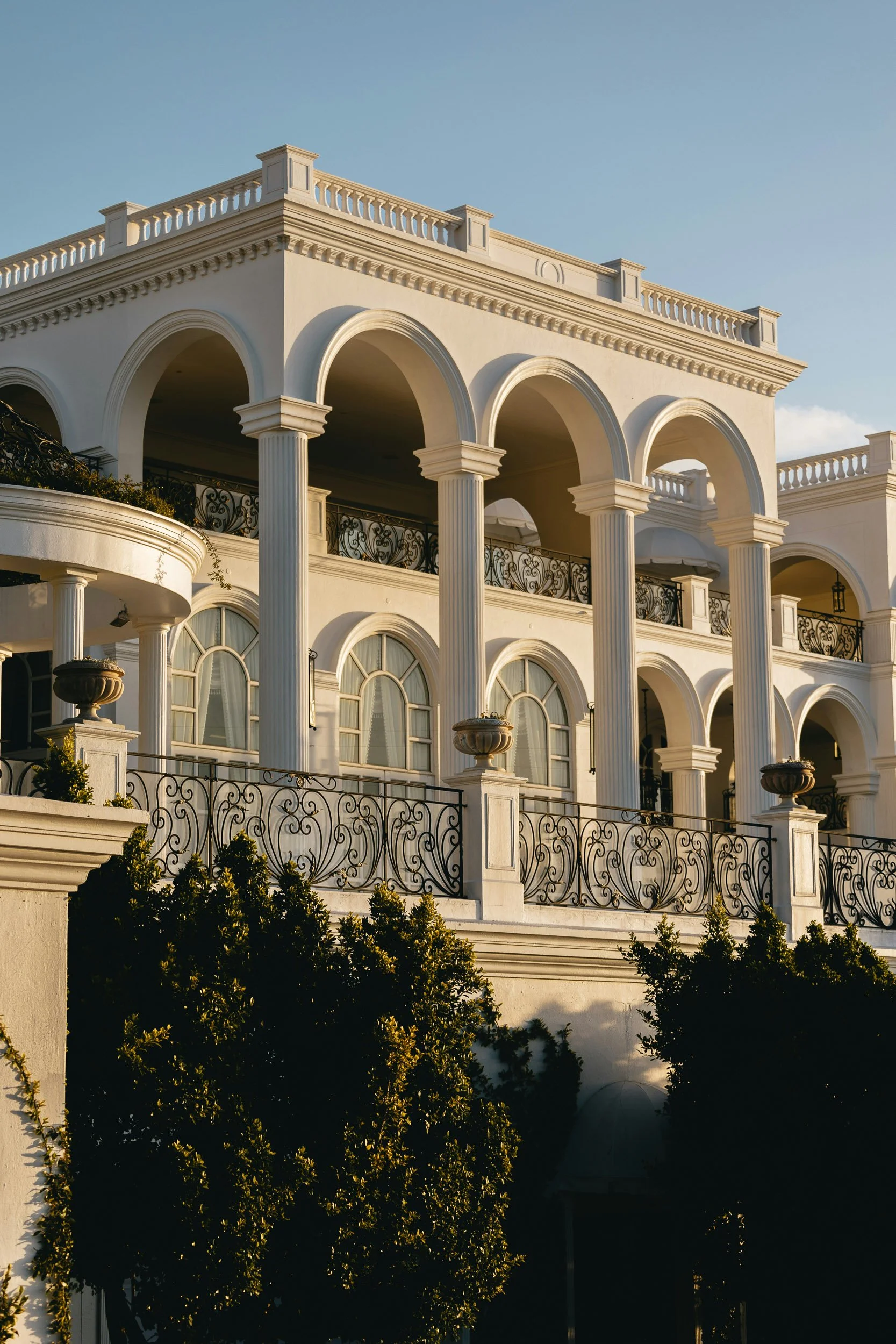A large elegant white building with classical architecture, featuring arched windows, columns, and ornate ironwork balconies, set against a clear blue sky.