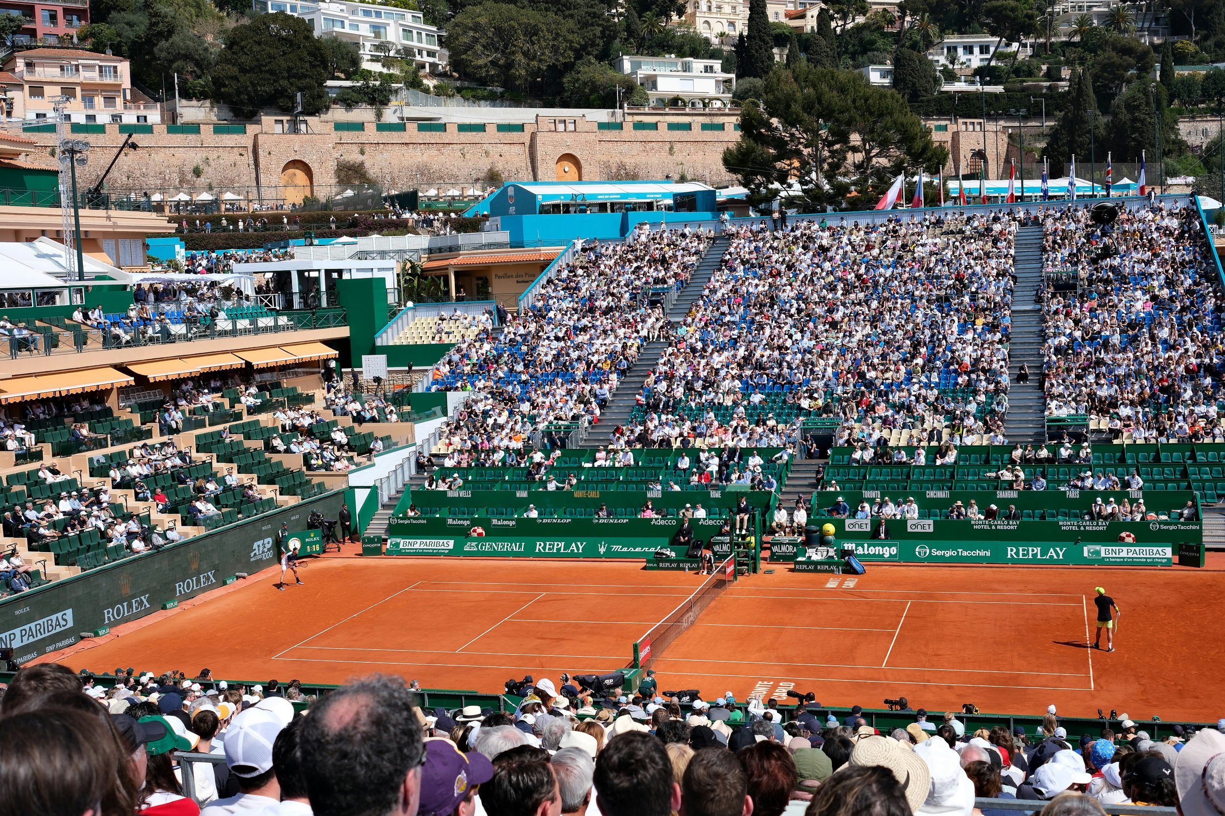 A crowded tennis stadium with many spectators watching a clay court tennis match, with a player at the baseline preparing to serve.
