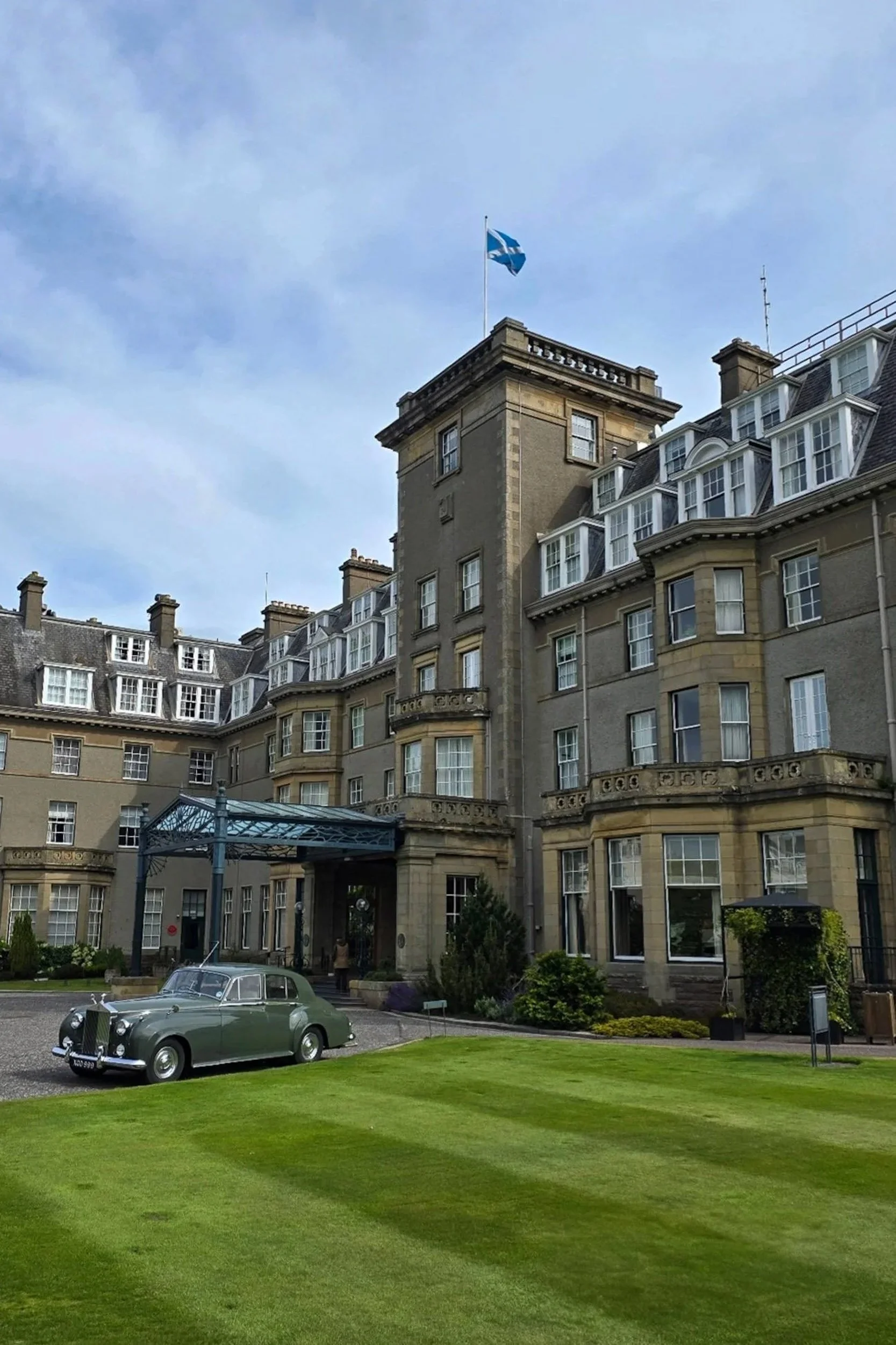 A vintage green car parked on a gravel driveway in front of a large, elegant hotel with multiple windows and a flag flying atop. The hotel has a classical architectural style, with a landscaped garden and a well-maintained lawn in the foreground.