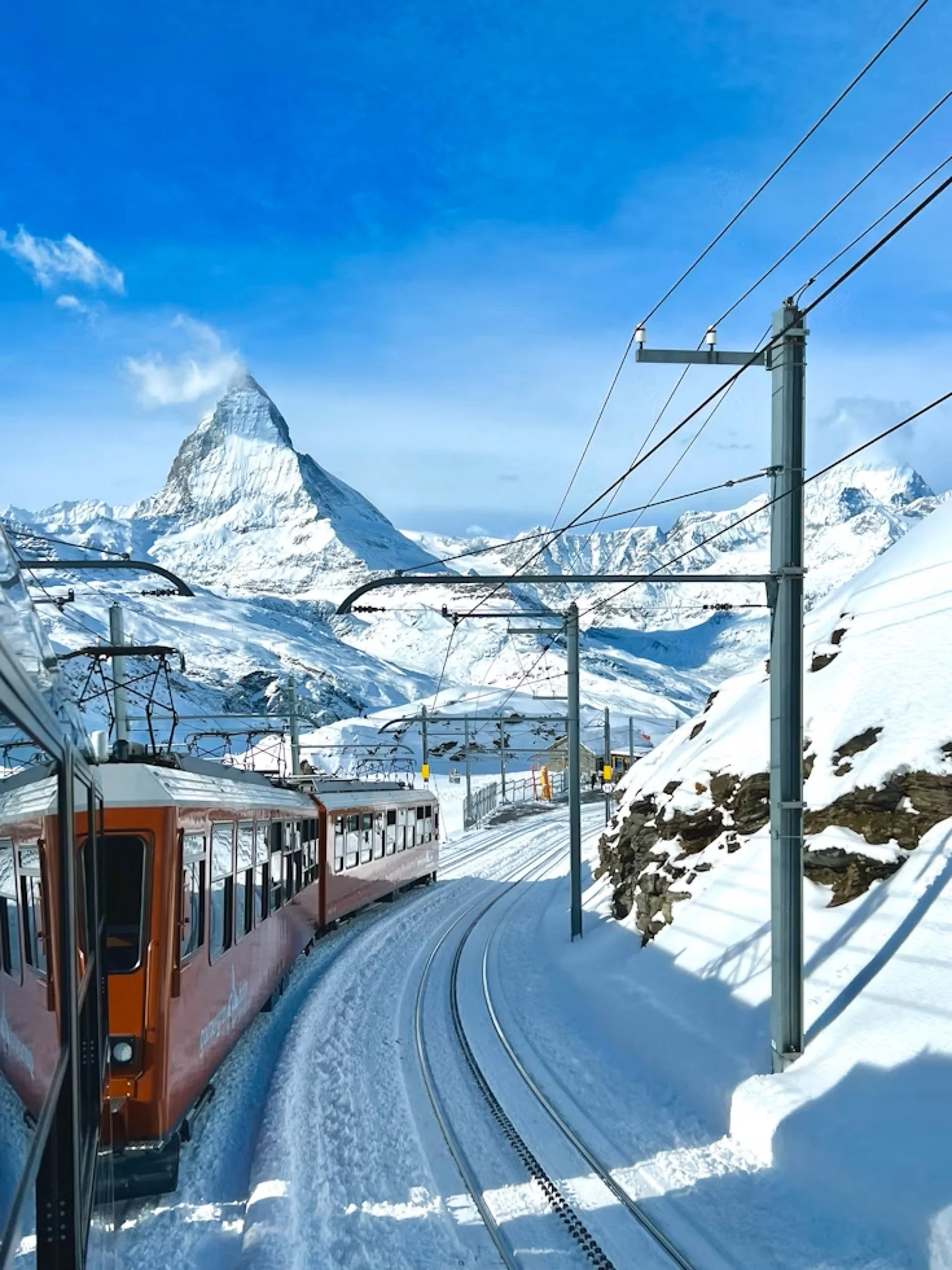 A ski train traveling through snowy mountains under a clear blue sky, with a prominent mountain peak in the background.
