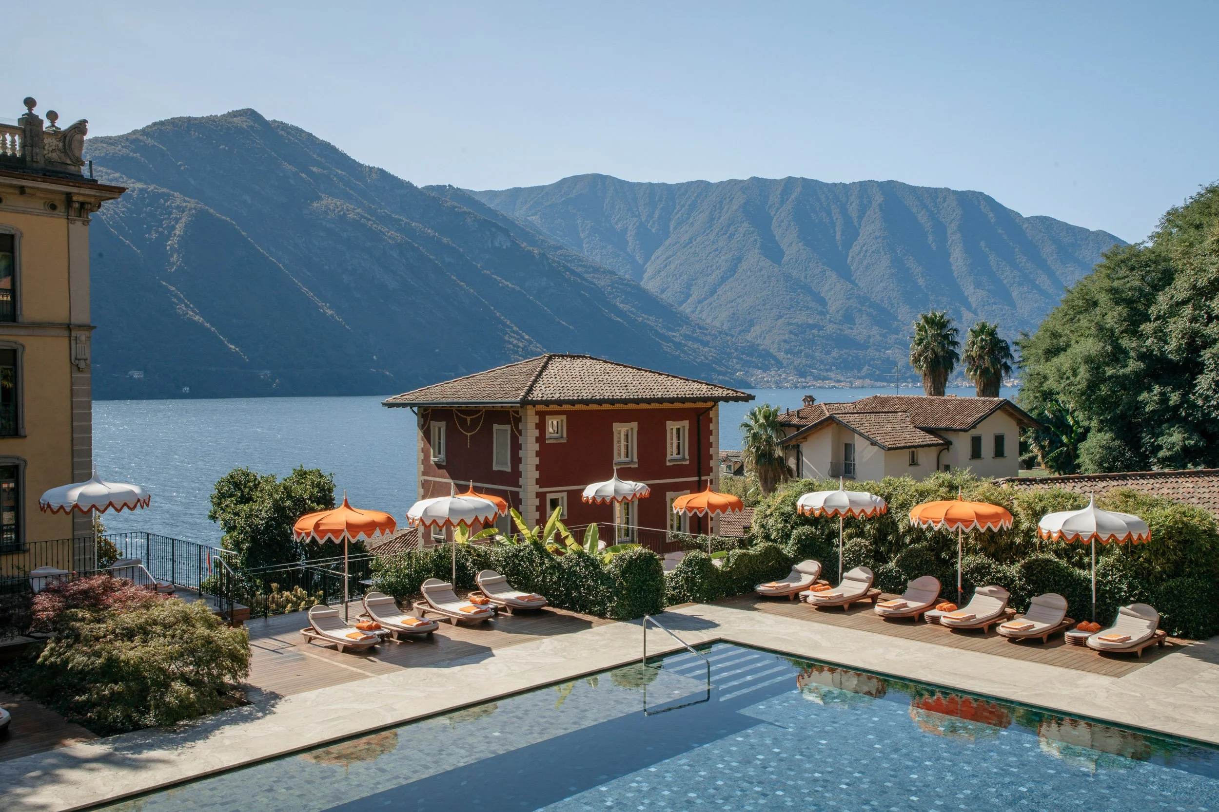 A scenic view of a lake surrounded by mountains, with a pool and lounge chairs in the foreground and umbrellas for shade.