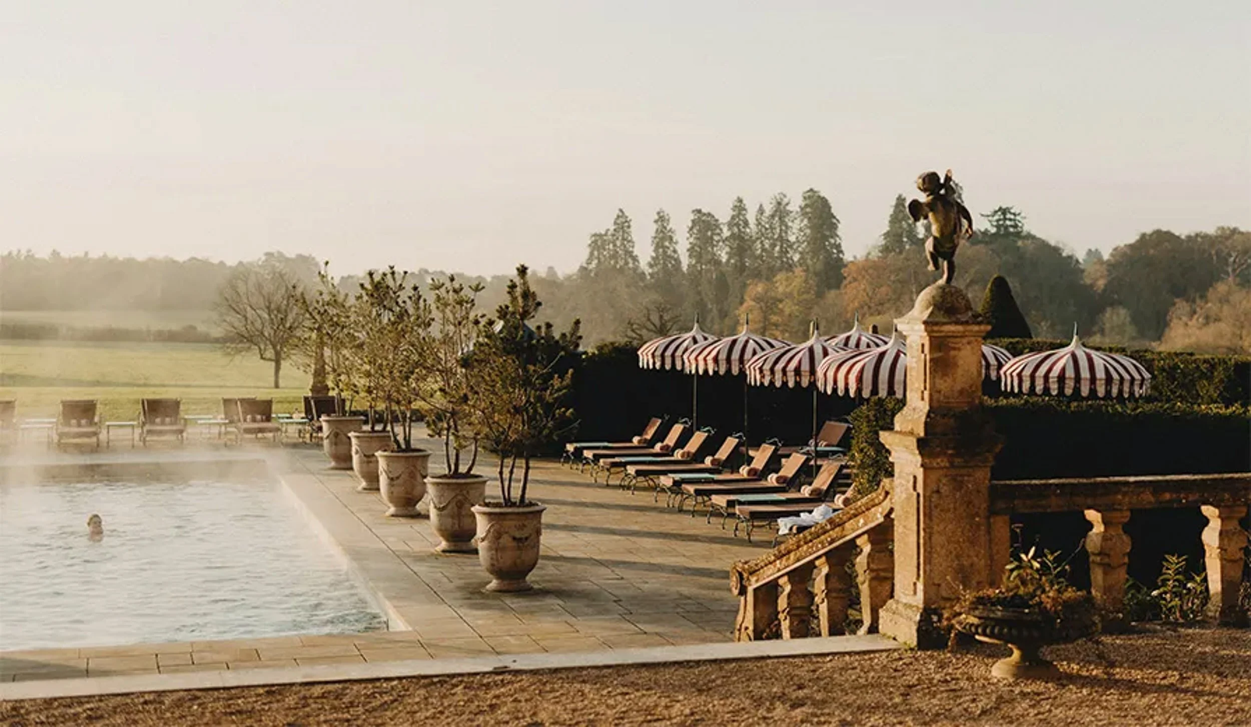 A luxurious outdoor swimming pool area with a person swimming, lined with potted trees, lounge chairs, and striped umbrellas. In the background, a statue of a dog with a bird on its back is situated on a stone pedestal, with trees and a clear sky visible.