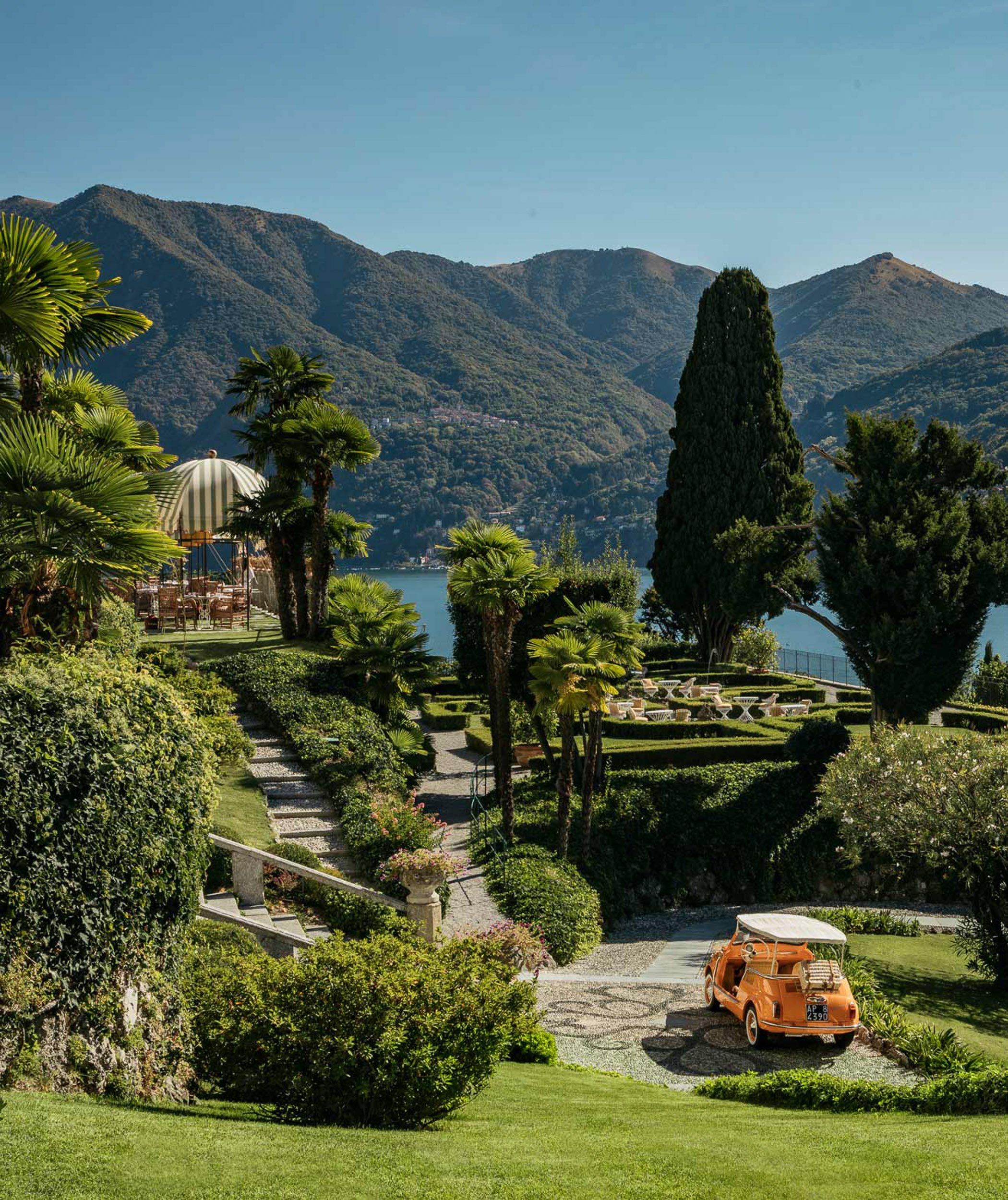 A scenic view of a well-maintained garden with a vintage orange car parked on a gravel driveway, surrounded by lush greenery, palm trees, and mountains in the background under a clear blue sky.