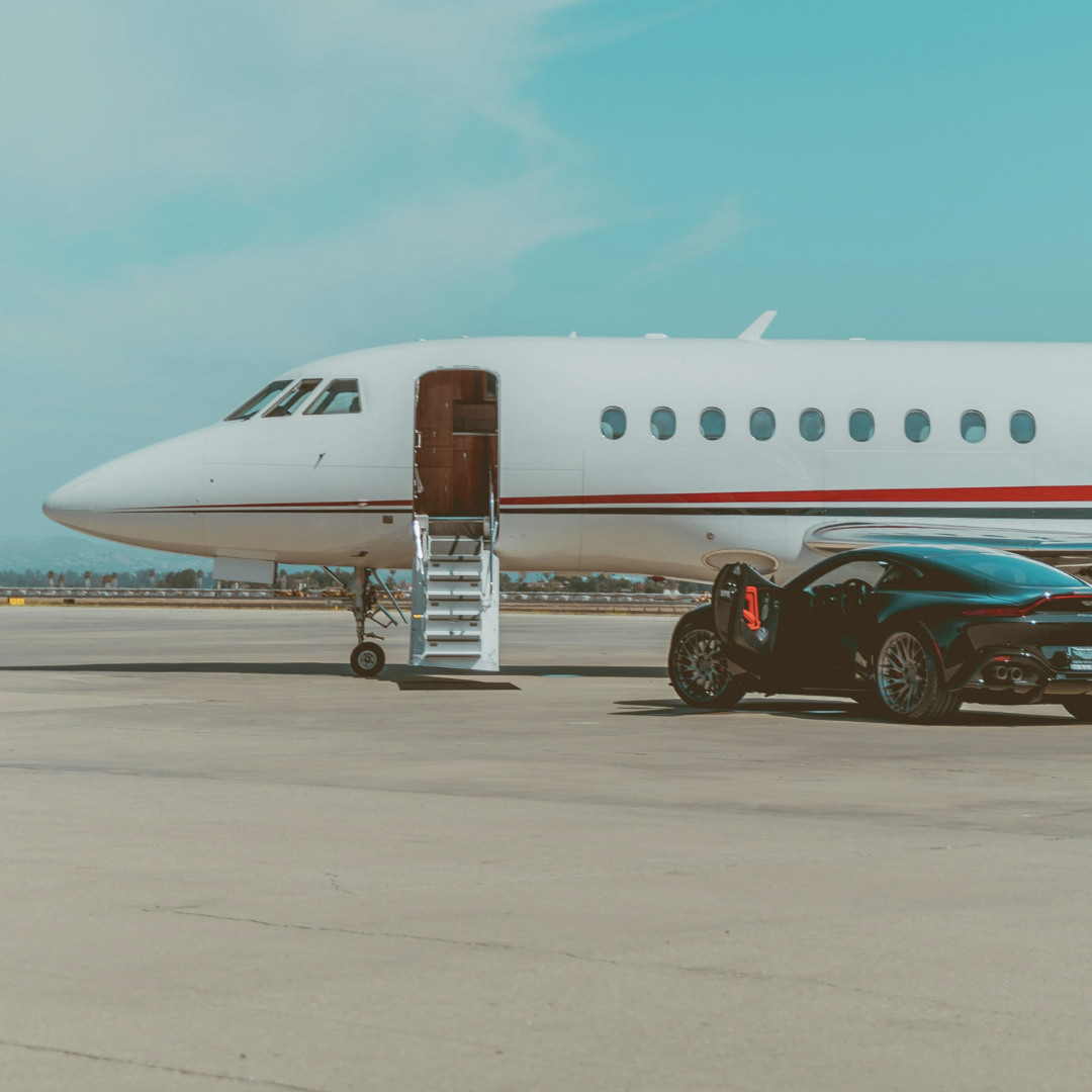 A white private jet with an open door and stairs on an airport tarmac, next to a black sports car.