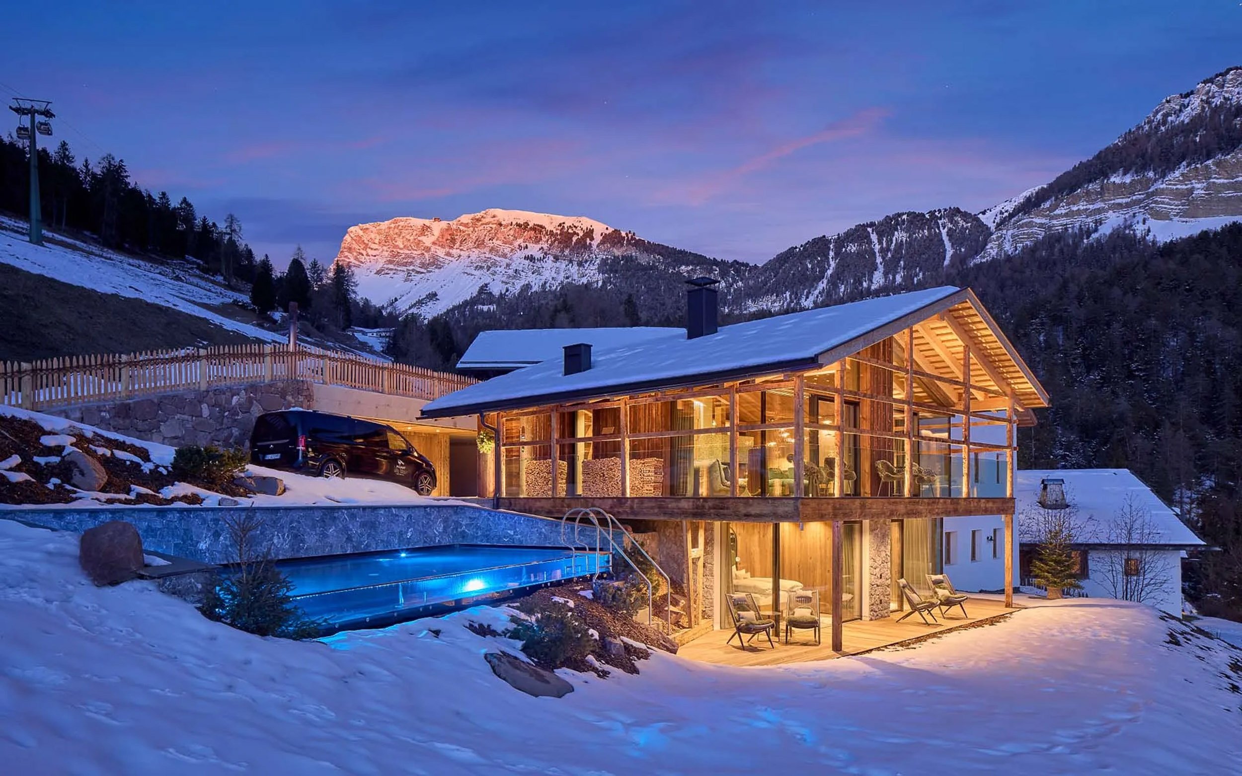 A modern wooden house with large glass windows and a sloped roof is illuminated from within, set amidst snowy mountains at dusk, with a mountain in the background bathed in pink and orange sunset light, and a small outdoor pool with blue lighting in the foreground.