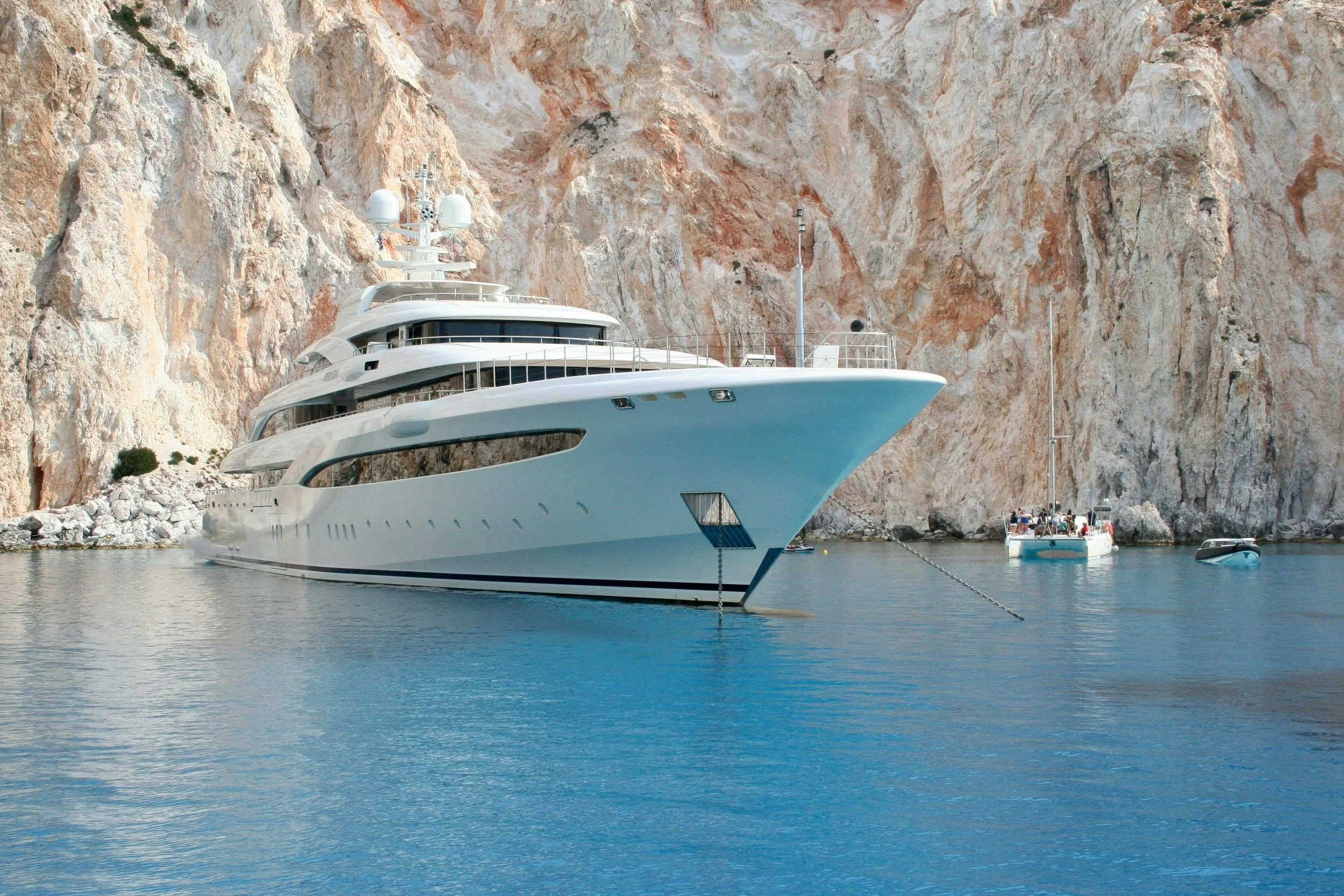 A large white luxury yacht docked in a calm harbor with rocky cliffs in the background. There are small boats with people on the water near the yacht.