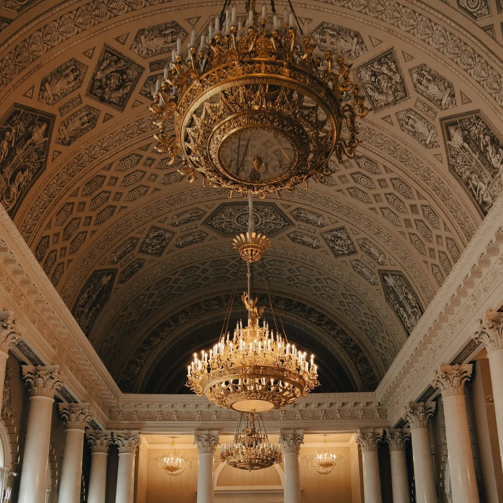 Interior view of a grand hall with a high ornate ceiling, decorated with intricate patterns and frescoes, featuring multiple large, elaborate chandeliers hanging from the ceiling.