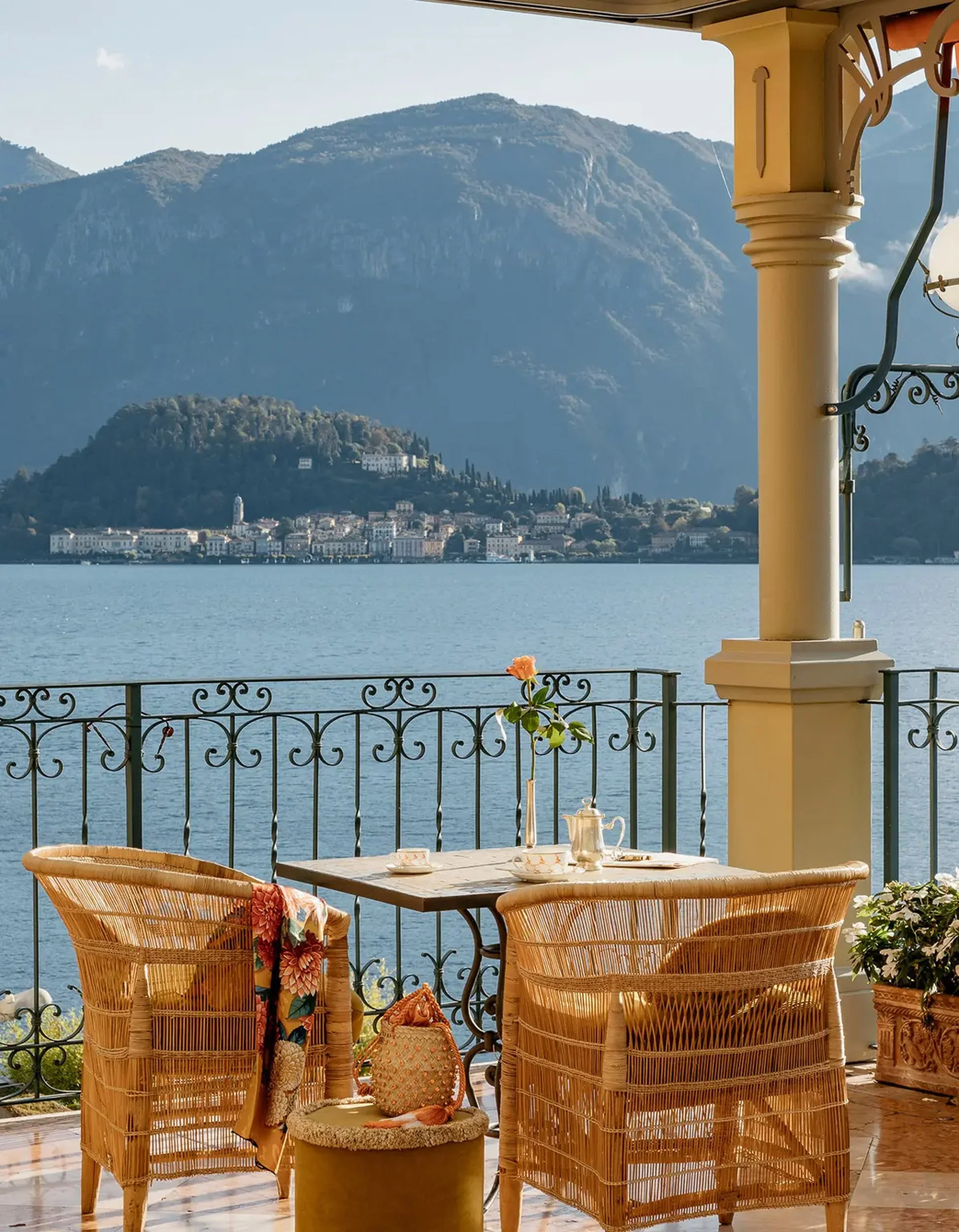 A balcony overlooking a lake with mountains in the background. The balcony has a table set with teapot, cups, and a flower in a pot. There are wicker chairs and a potted plant.