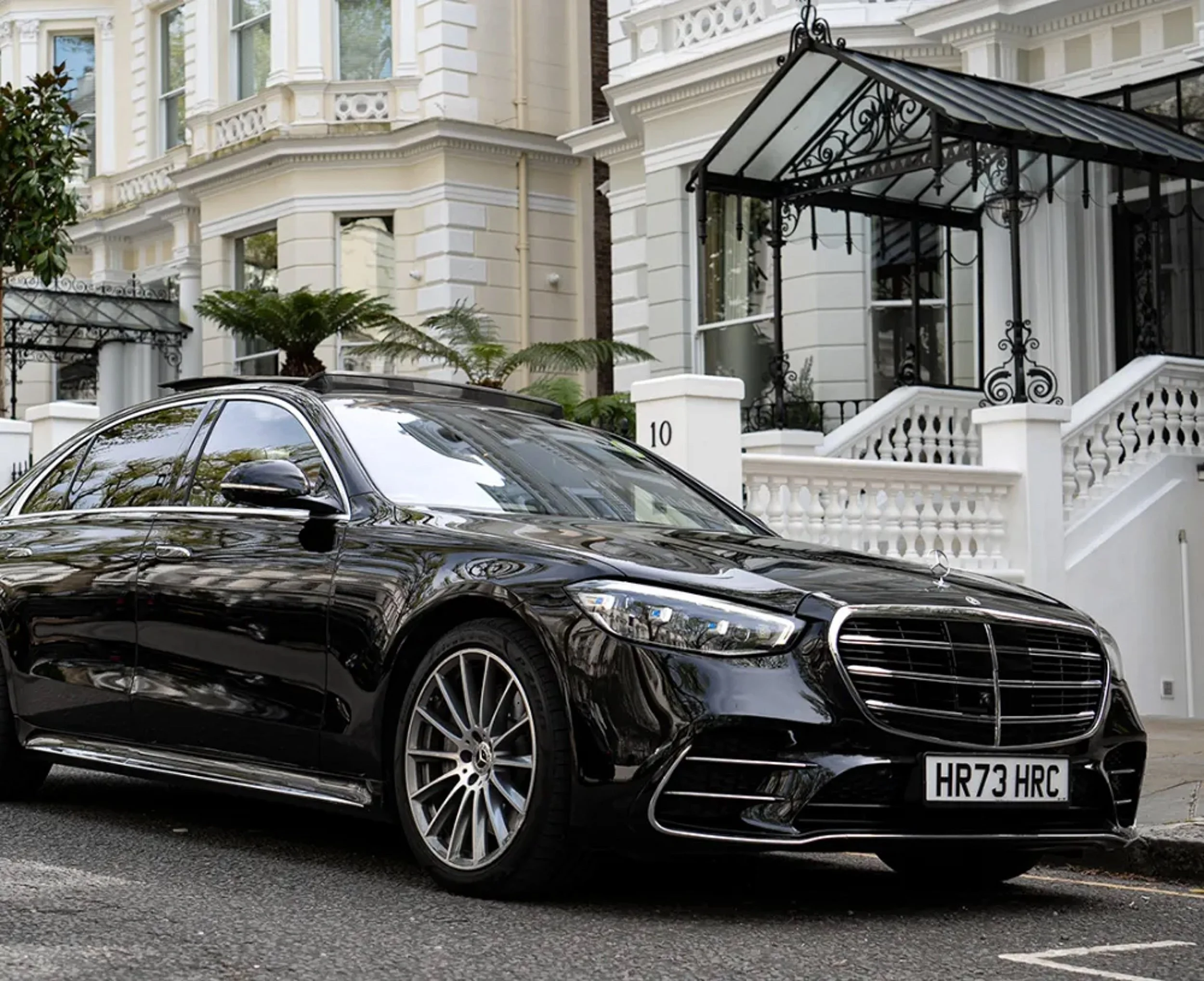 A black luxury Mercedes-Benz parked in front of a white house with ornate architectural details and security features, including a wrought iron canopy and a white residential gate. The car's license plate reads HR73 HRC.