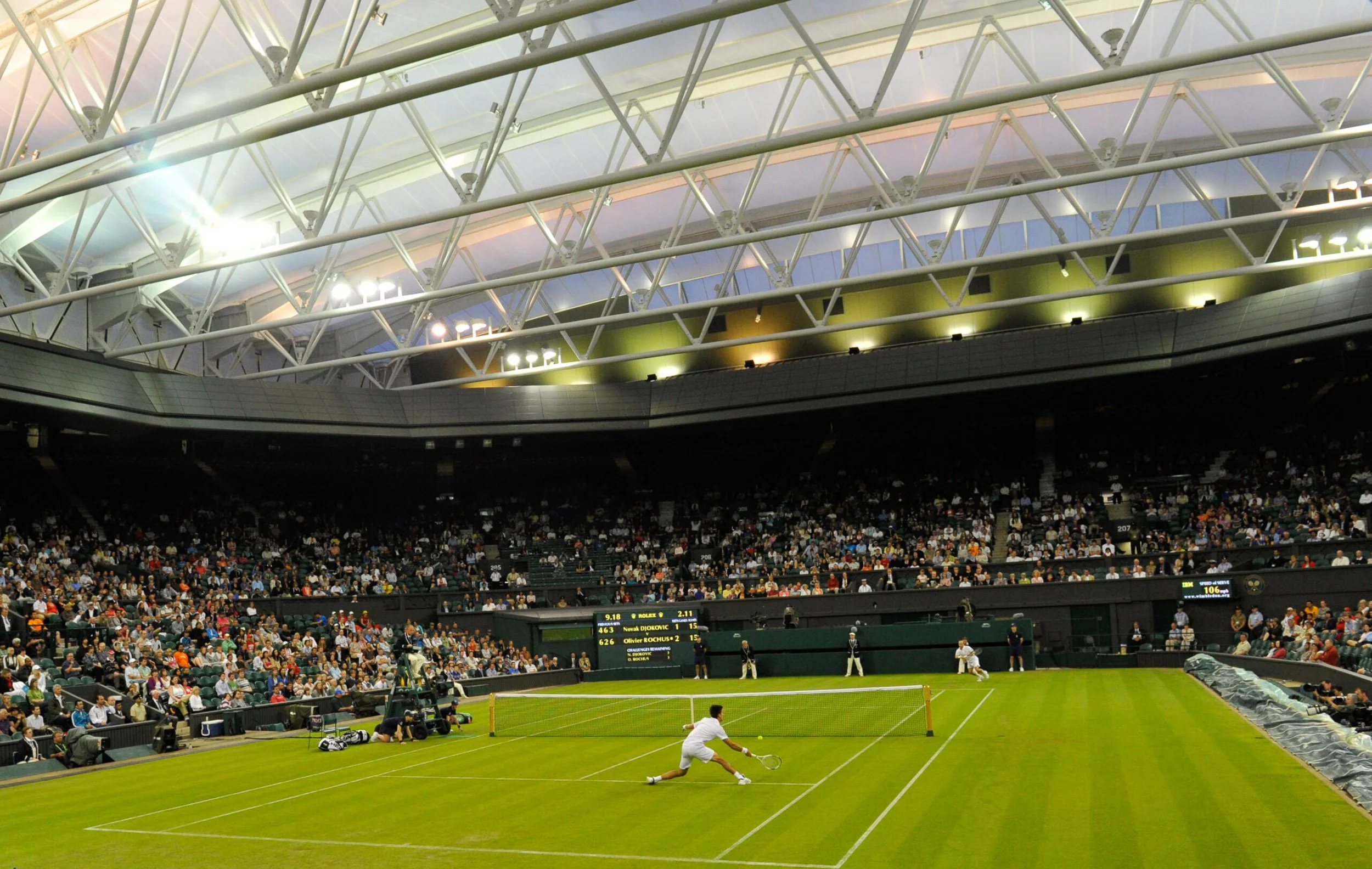 Tennis player in white uniform lunging to hit a tennis ball on a green court inside a large stadium with a metal roof and a crowd of spectators.