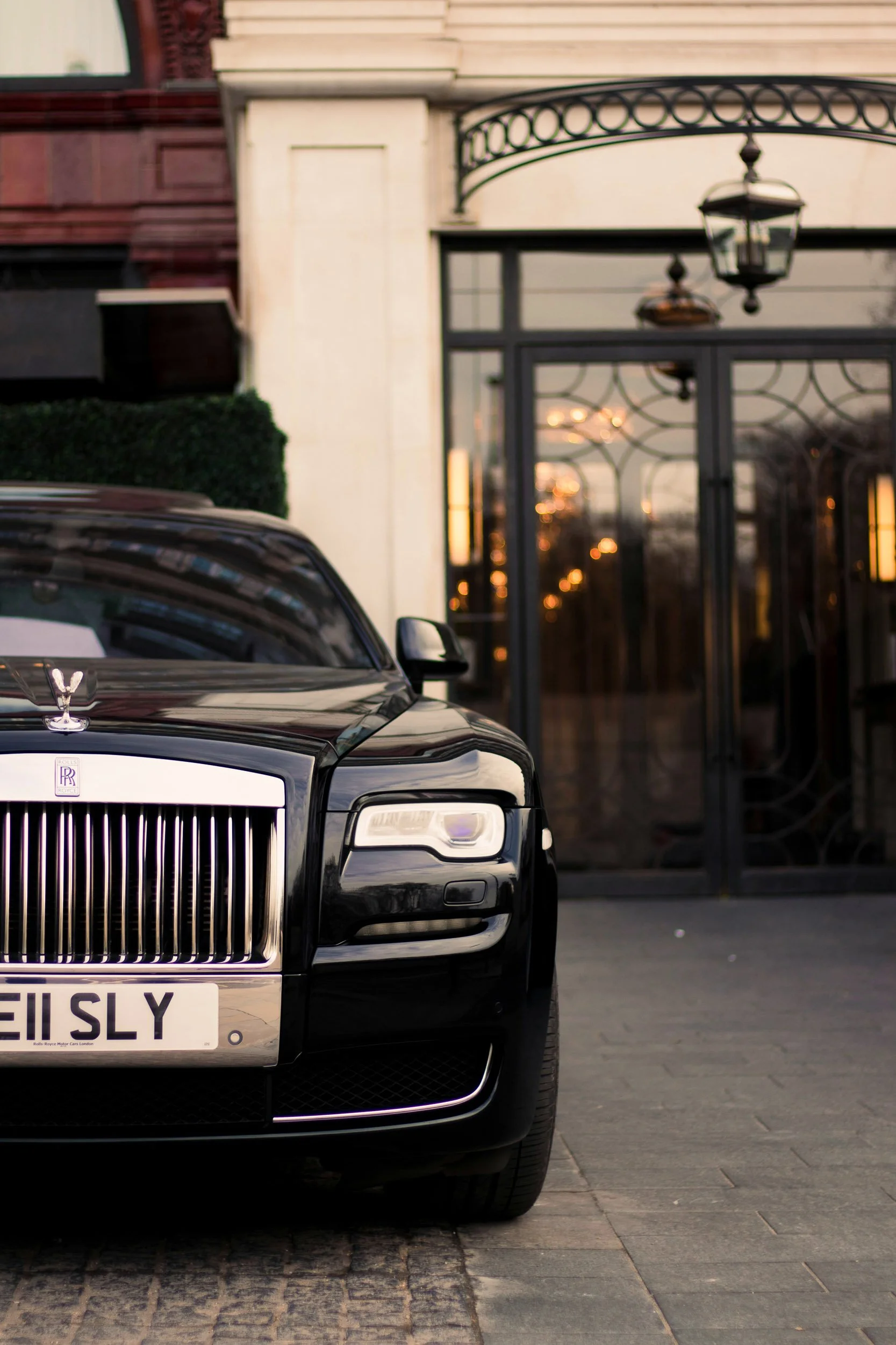 Close-up of a black Rolls-Royce luxury car parked in front of a decorative gate and building; focus on the front grille, hood ornament, and part of the license plate.