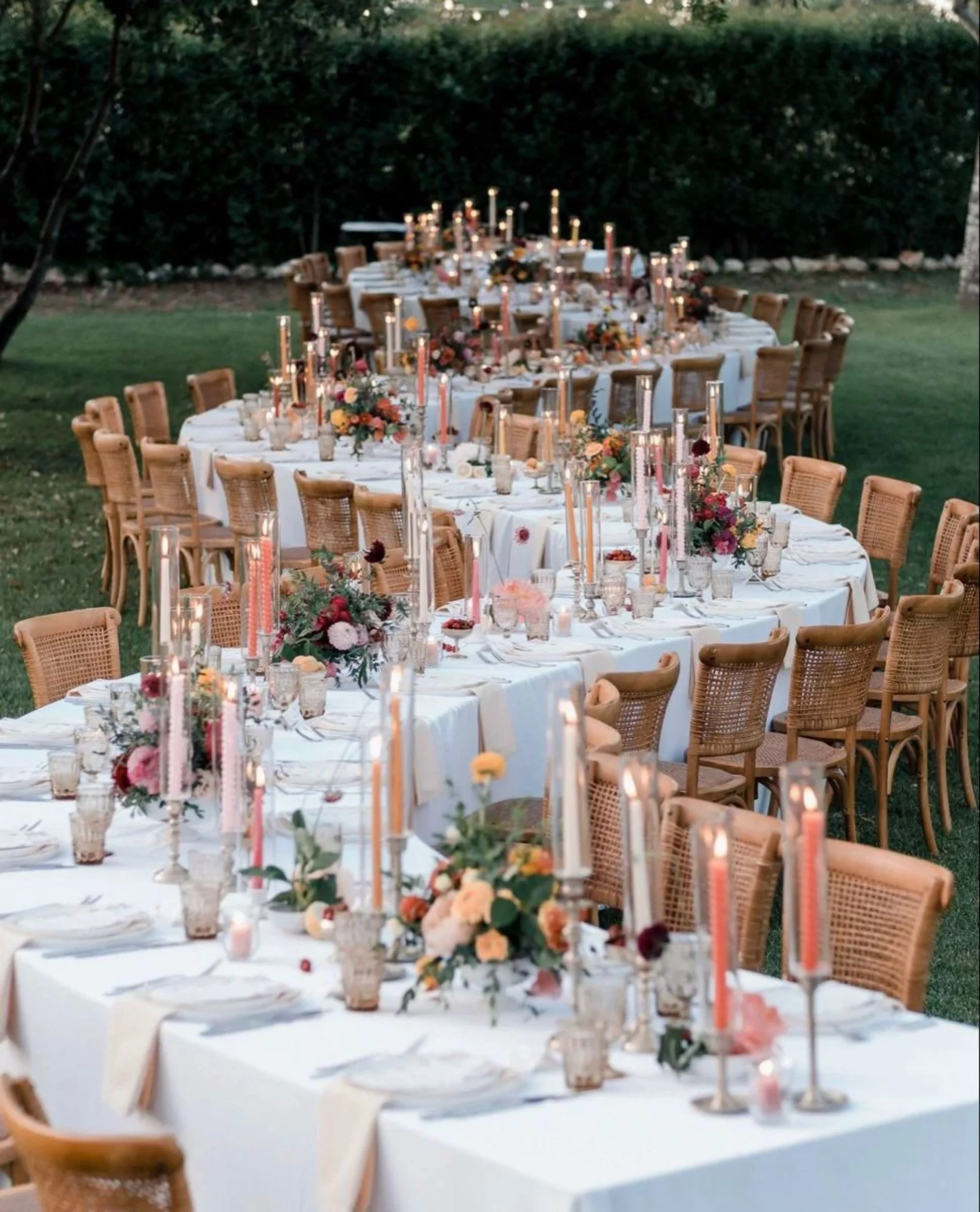 Round outdoor dining tables decorated with floral arrangements and tall candles in a garden setting, prepared for a special event or celebration.