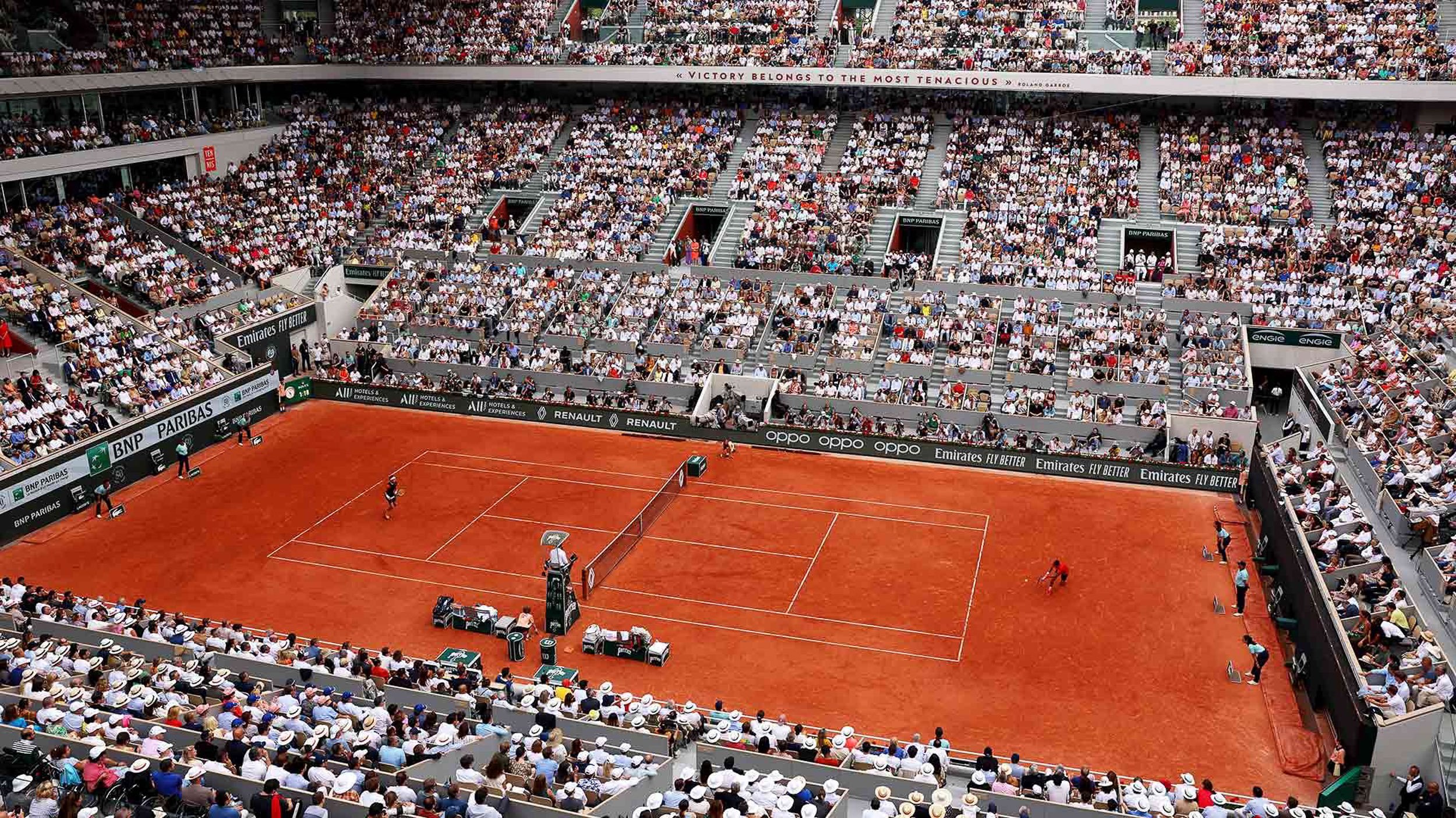A crowded tennis stadium filled with spectators watching players on a clay court, with visible sponsor banners.