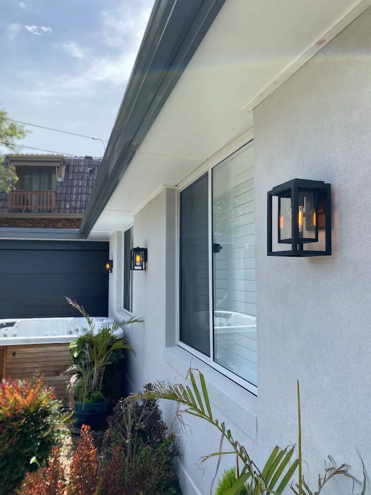 Exterior wall of a house with two black wall-mounted lantern-style lights, a window with white shutters, and a garden with various plants and a hot tub in the background.