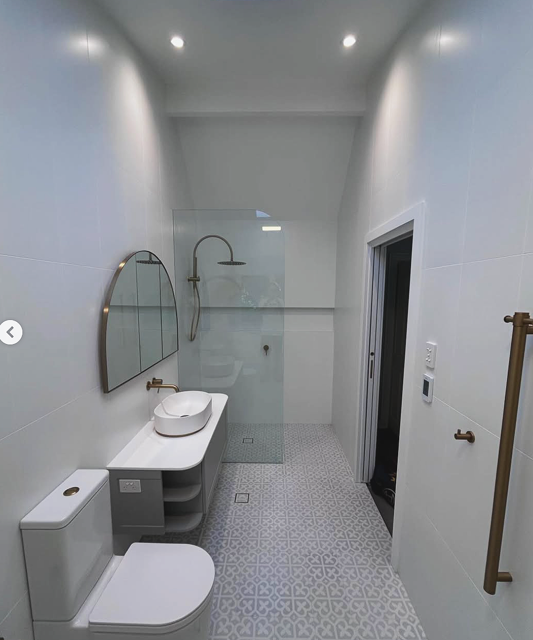 A modern bathroom with white walls and patterned floor tiles. It has a toilet, a floating vanity with a vessel sink, a rounded mirror, a glass-enclosed walk-in shower with a rainfall showerhead, and a door leading to another room. The bathroom features gold fixtures and recessed ceiling lights.