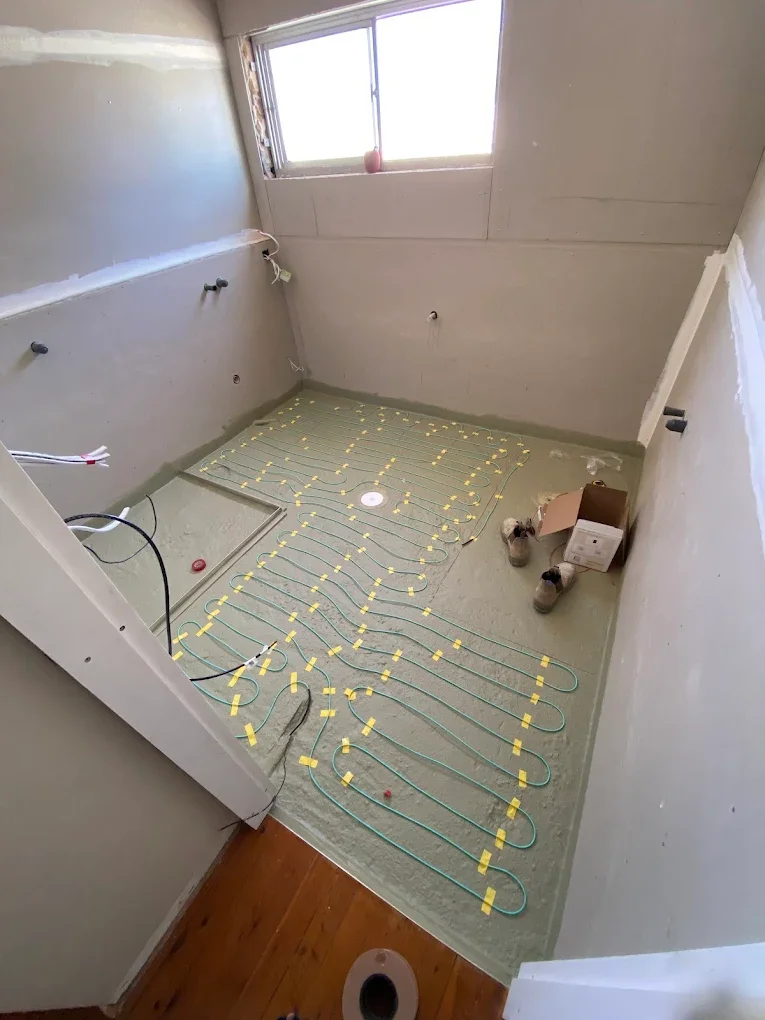 Bathroom under renovation with underfloor radiant heating pipes installed on the floor, yellow tape markings, and exposed pipes and wires on the walls. There's a window at the far end and some tools and boxes on the floor.