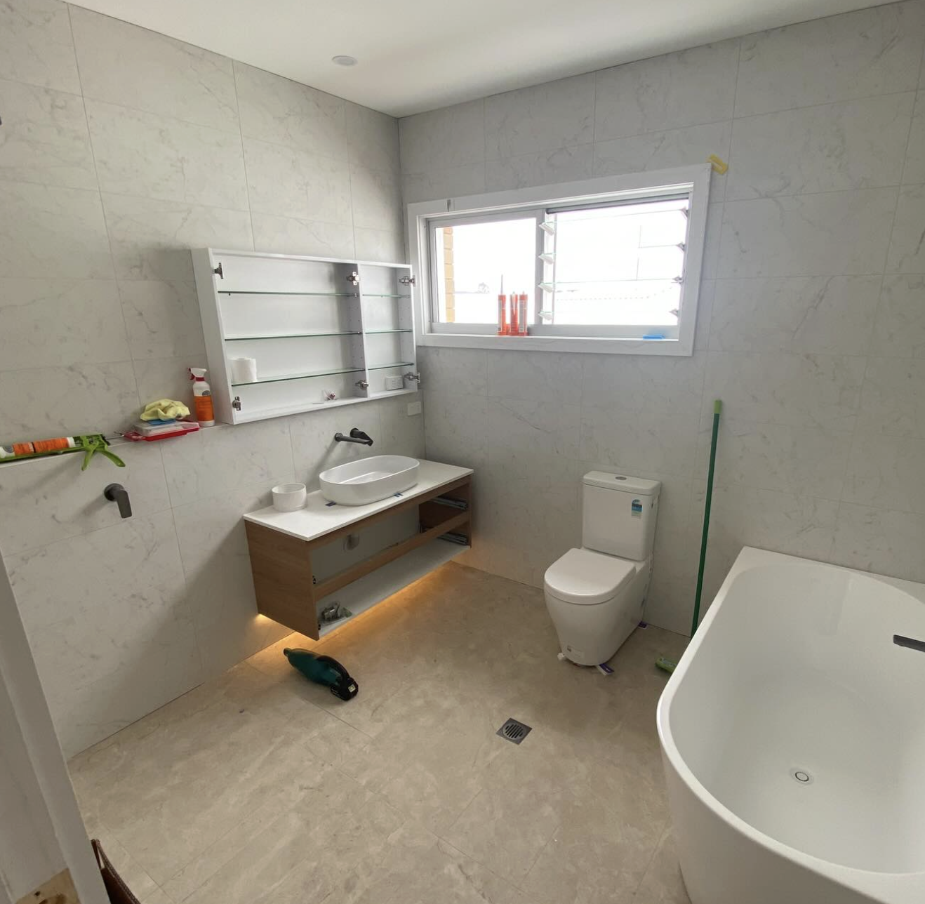 A modern bathroom with a white tub, toilet, and a minimalistic floating vanity with a vessel sink. There is a window with security bars, a wall-mounted glass shelf, and cleaning tools on the floor.