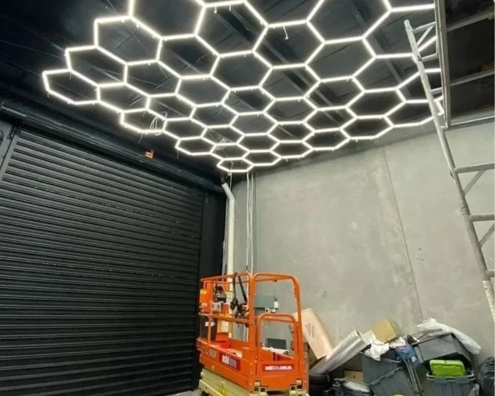 Interior of a building under construction, showing LED hexagonal light fixtures on the ceiling, a black rolling door, a scissor lift, and construction materials.
