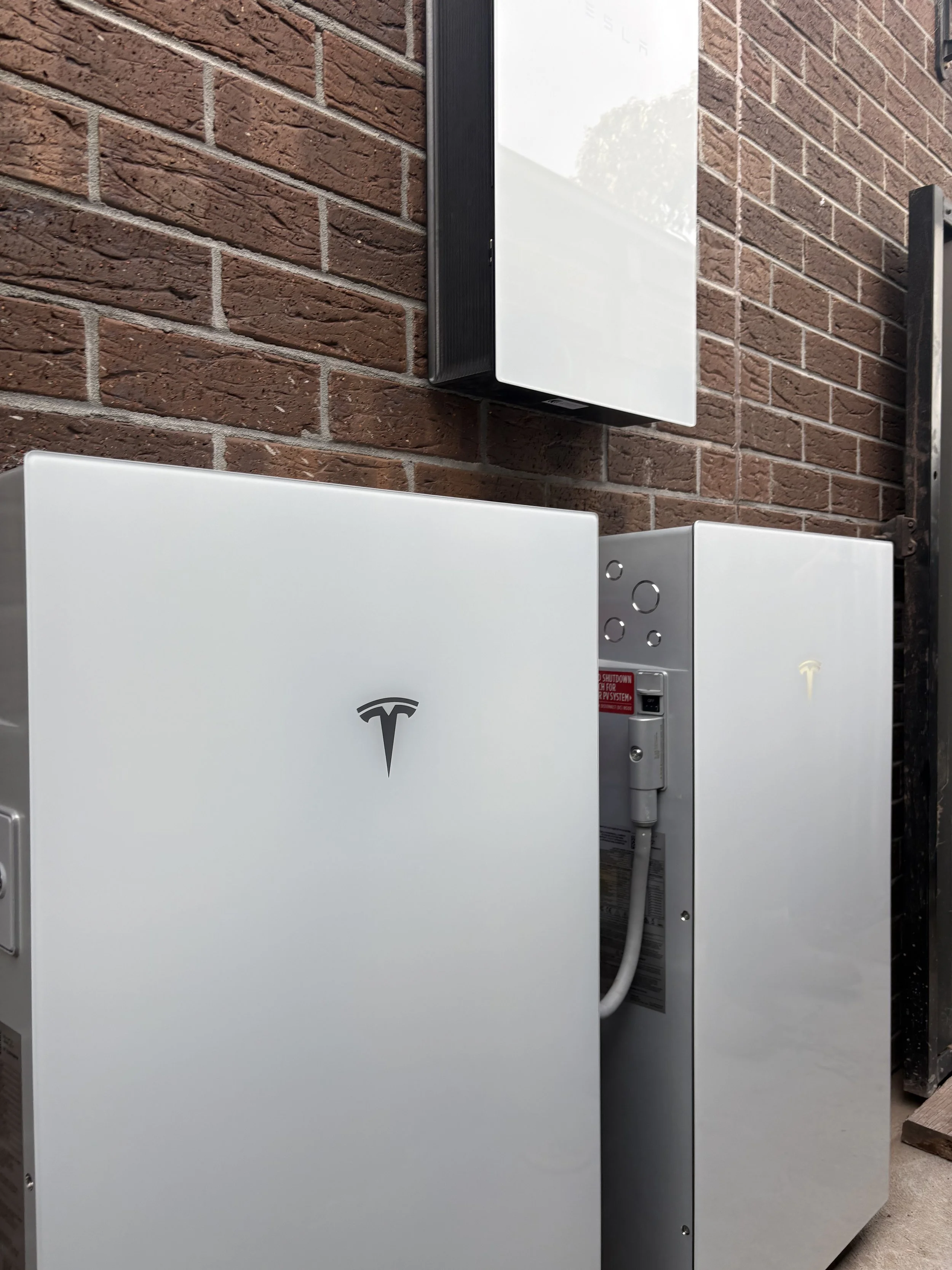 White Tesla Powerwall units mounted on a brick wall, with electrical conduits and a control panel.