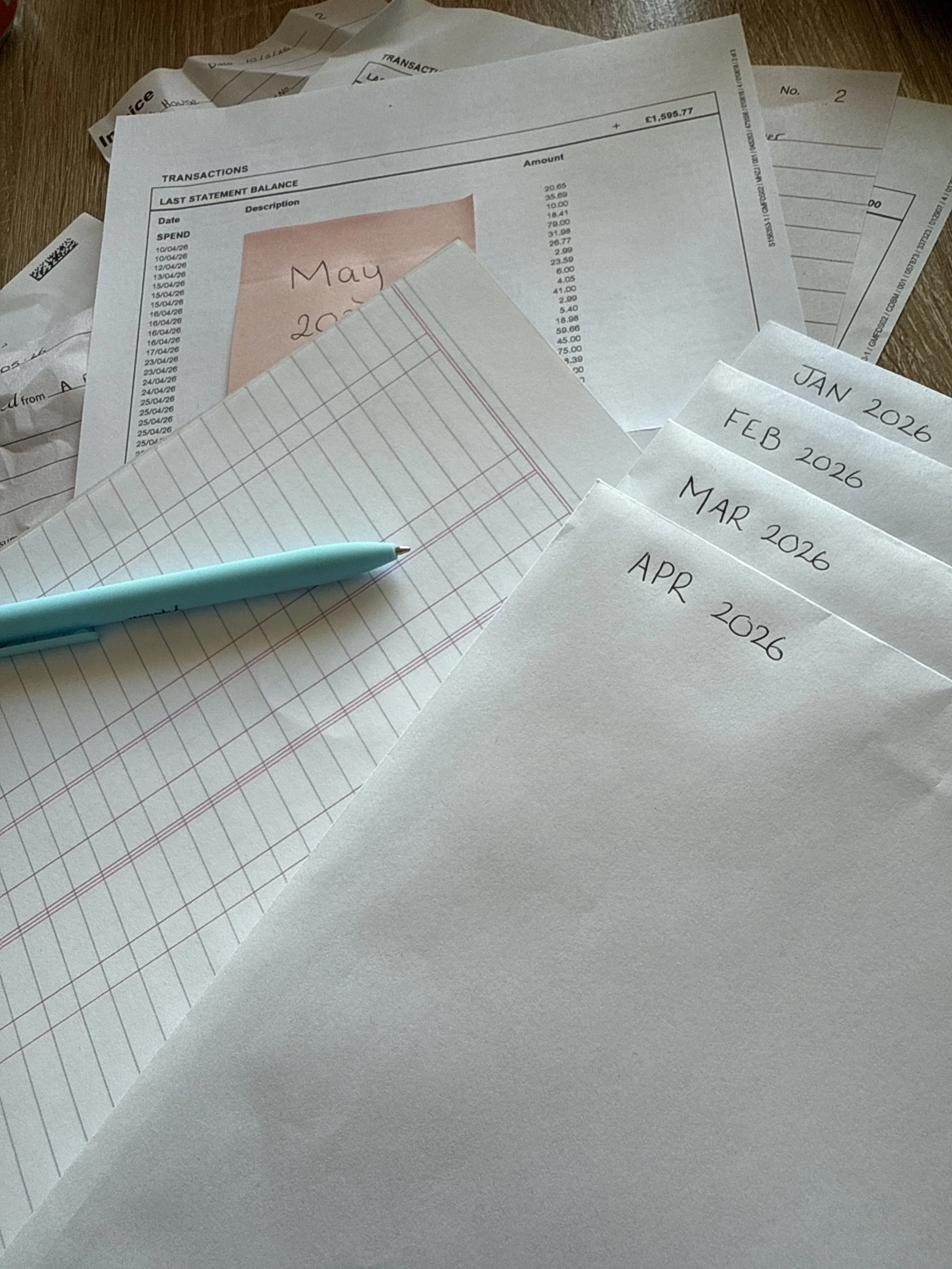 A cluttered desk with receipts, handwritten notes, a notepad, and a blue pen, covering months from February to May in 2026.