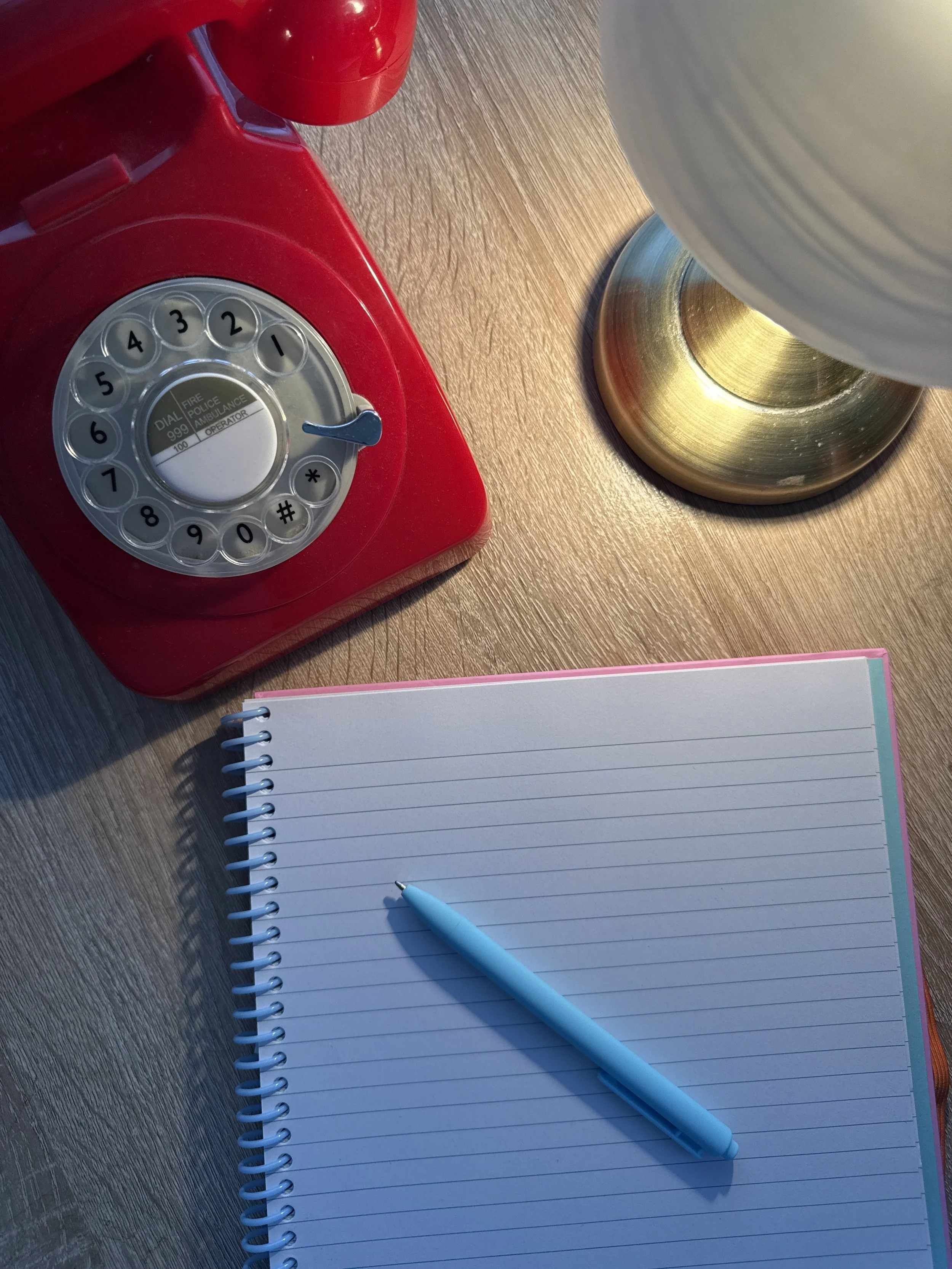 A red rotary phone, a spiral-bound notebook with lined paper, a light blue pen, and a brass-colored cup with a large white mug inside it on a wooden surface.