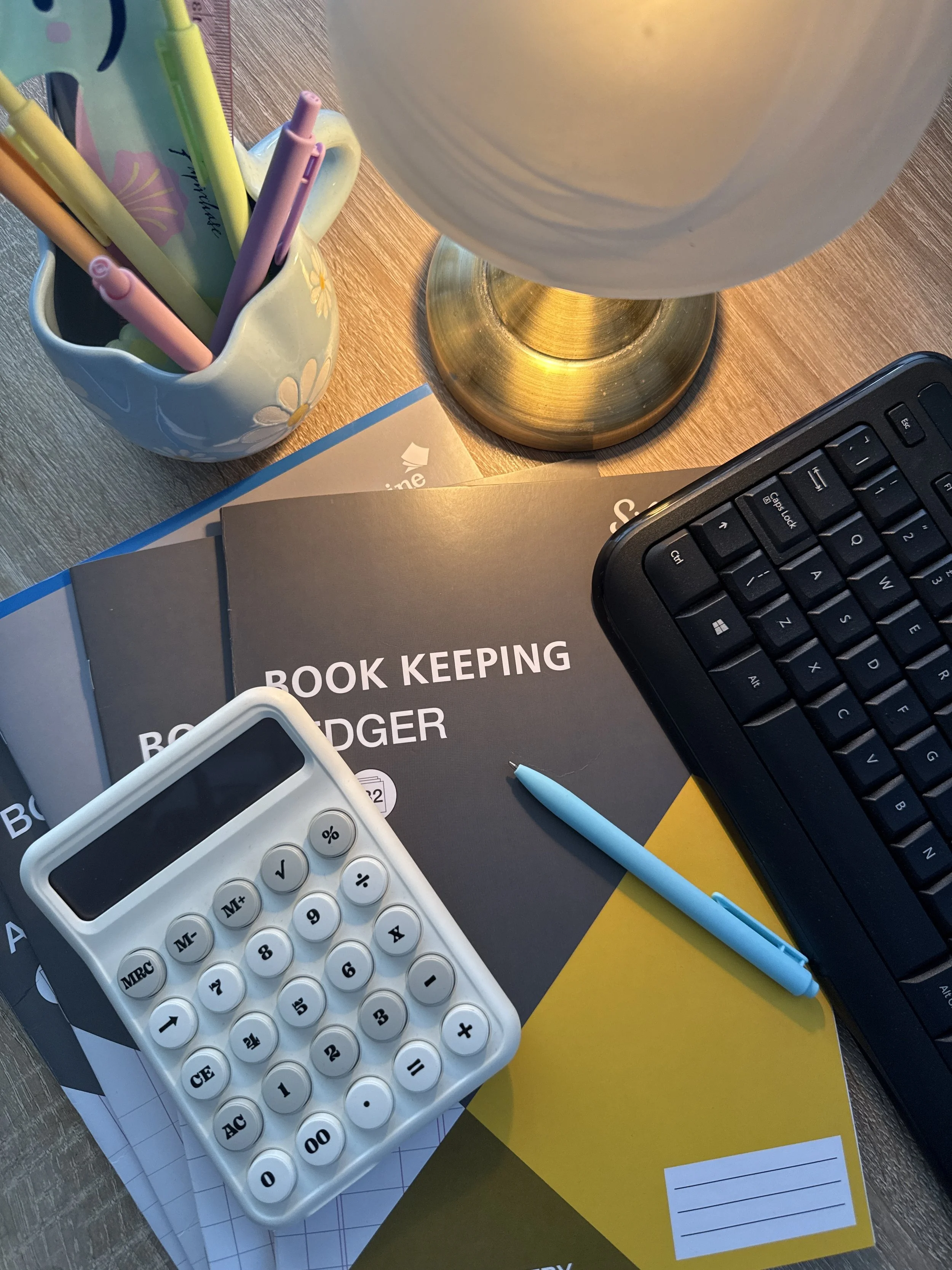 Desk with a lamp, a calculator, pens in a mug, a keyboard, a book titled 'Book Keeping,' and notebooks.