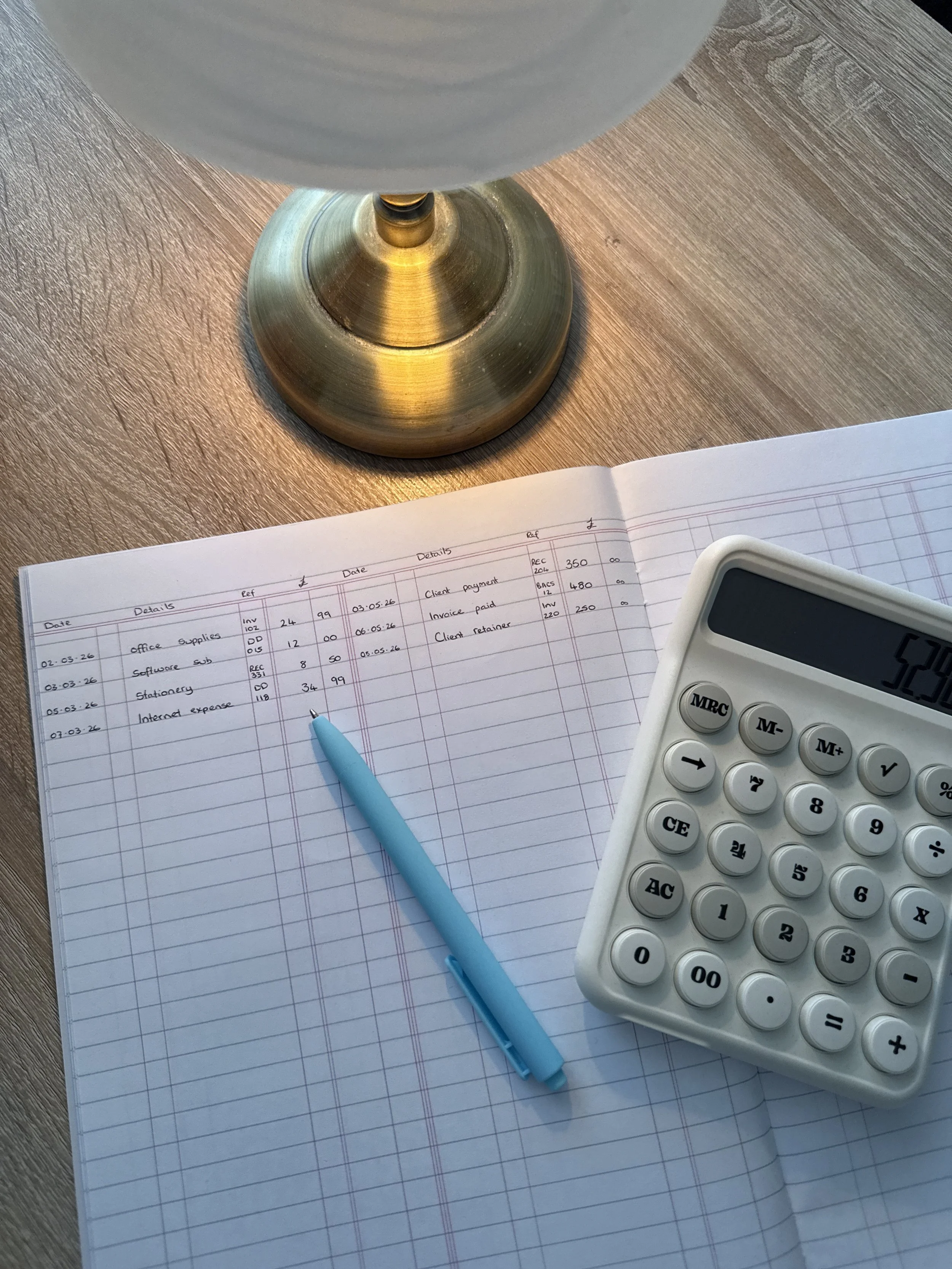 Close-up of a desk with a paper ledger containing financial entries, a blue pen, a white calculator, and a brass table lamp on a wooden surface.