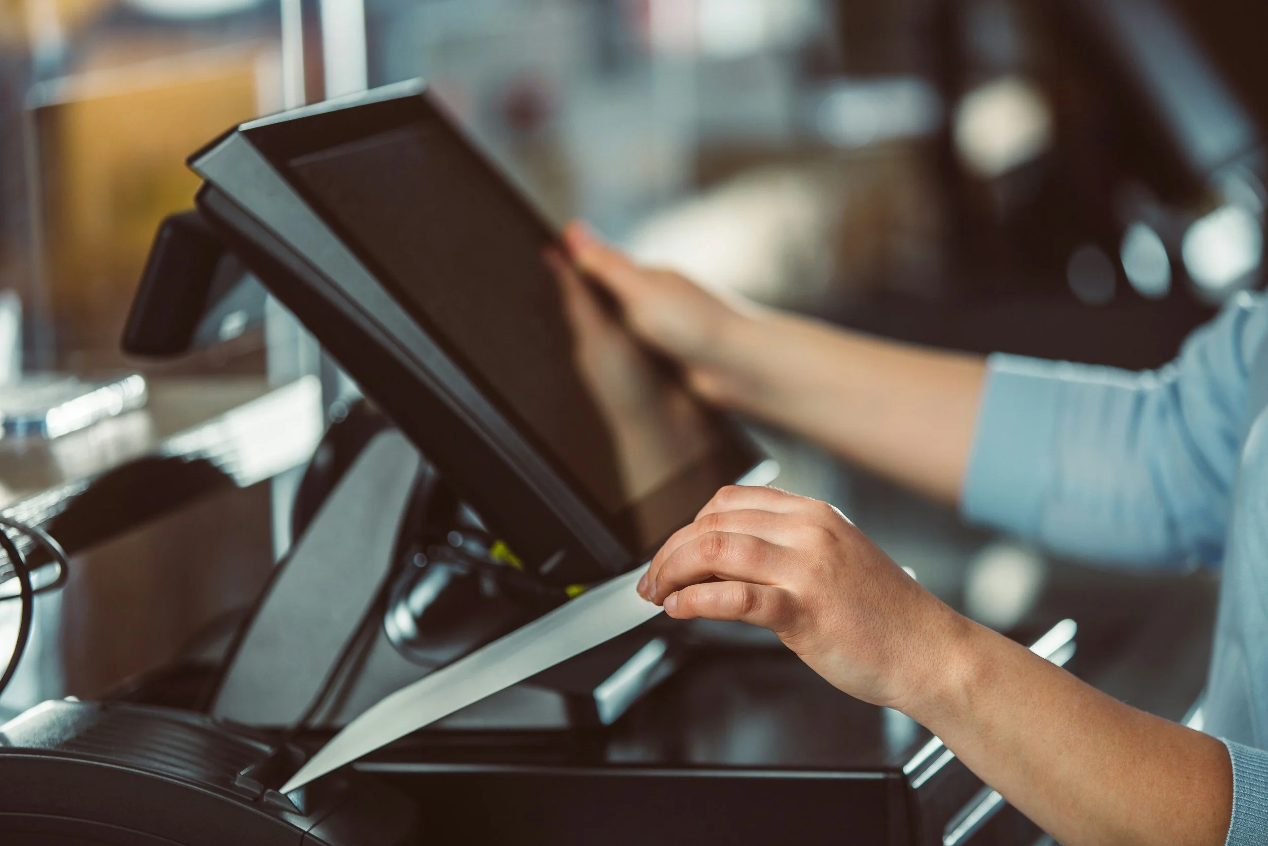 Close-up of a person's hands operating a point-of-sale touchscreen device, possibly a tablet or monitor, with a blurred background.