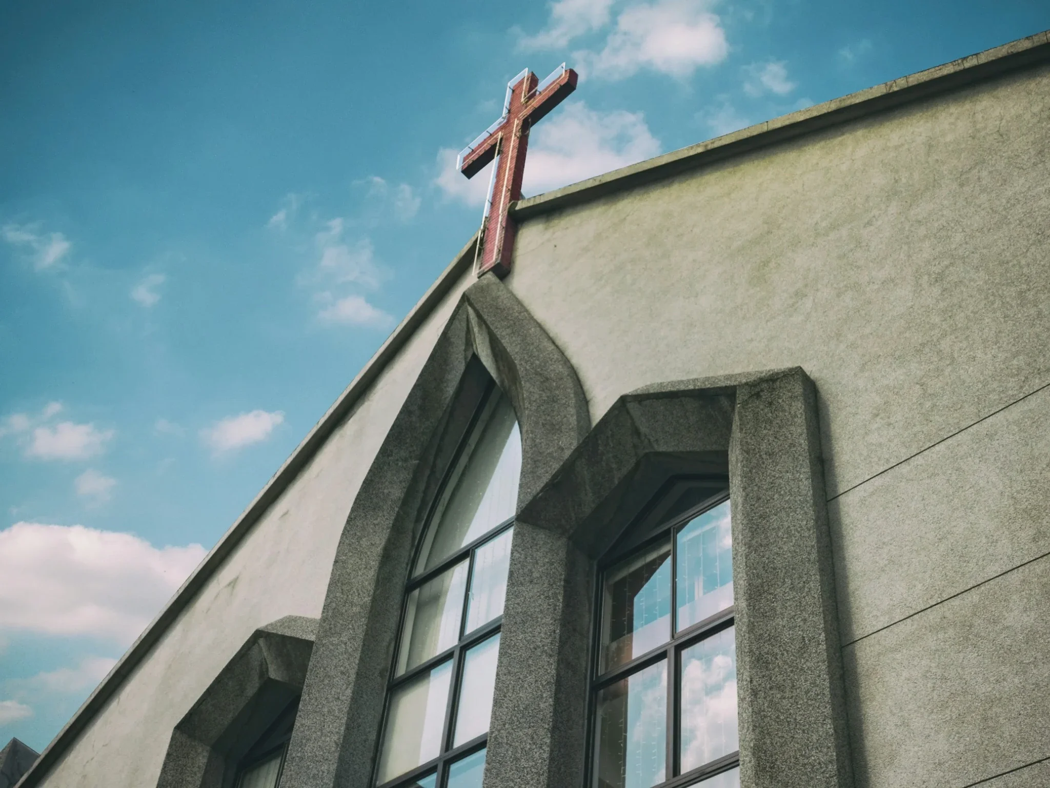 Close-up of a church steeple with a cross on top, attached to a modern building with large glass windows and stone accents, against a blue sky with clouds.