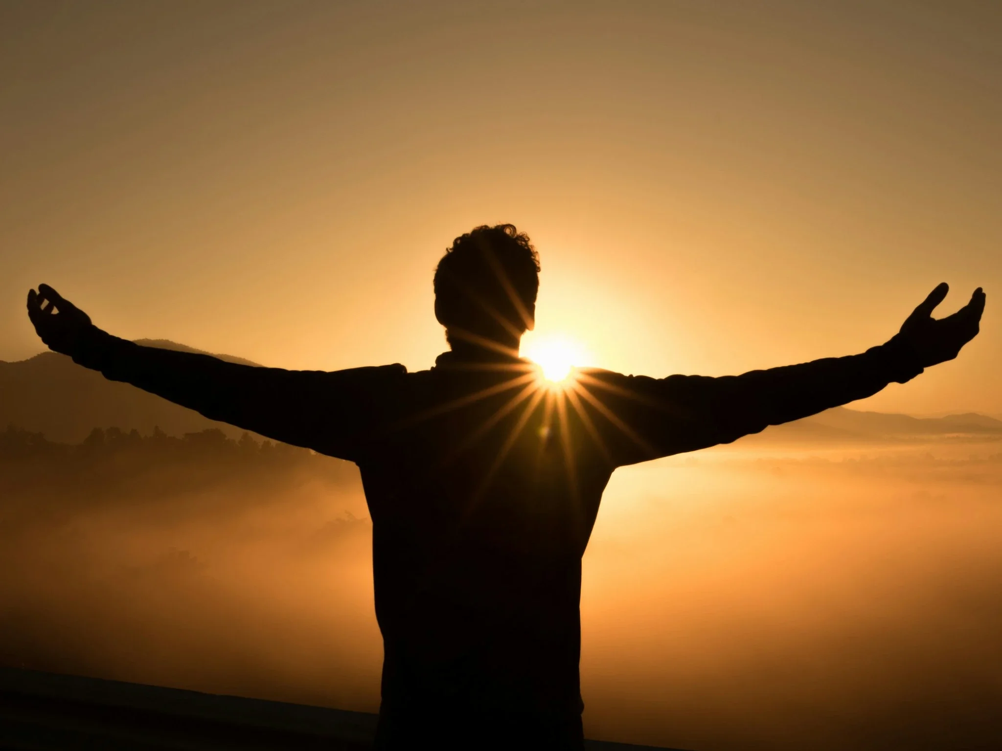 Silhouette of a person with arms extended outward against a sunset, with the sun shining through their hands, and a landscape in the distance.