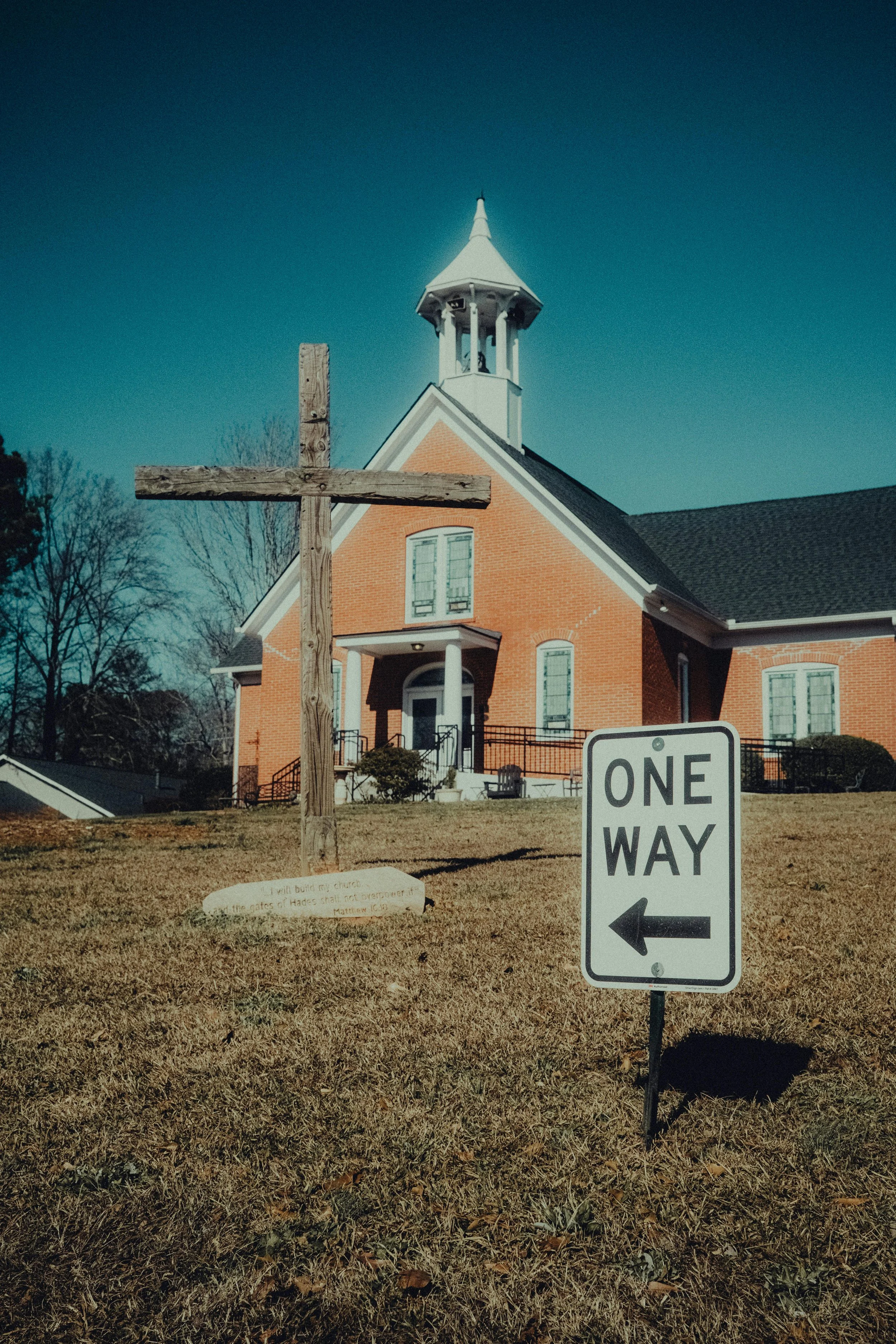 A red-brick church with a white steeple and a cross on a pole in the front lawn. There is a one-way sign pointing to the left and a wooden sign with a quote near the grass.