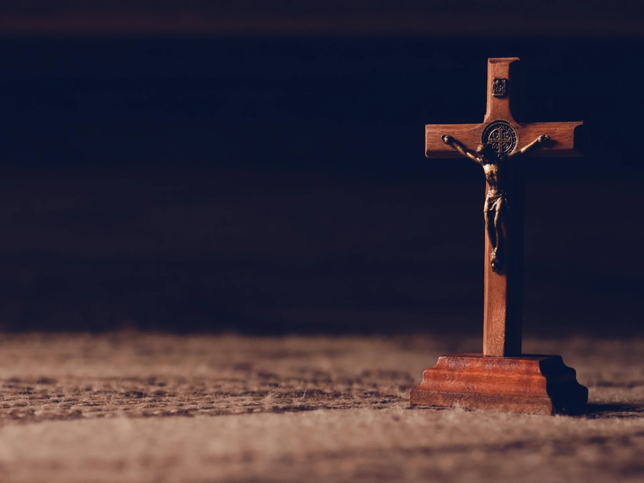 A wooden crucifix with Jesus Christ mounted on a stand, standing on a rough surface against a dark sky.