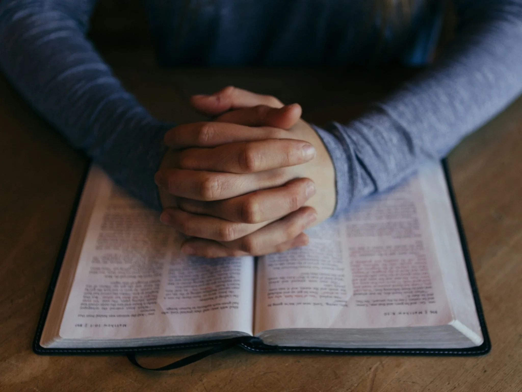 Open Bible resting on a table with hands clasped in prayer above it.