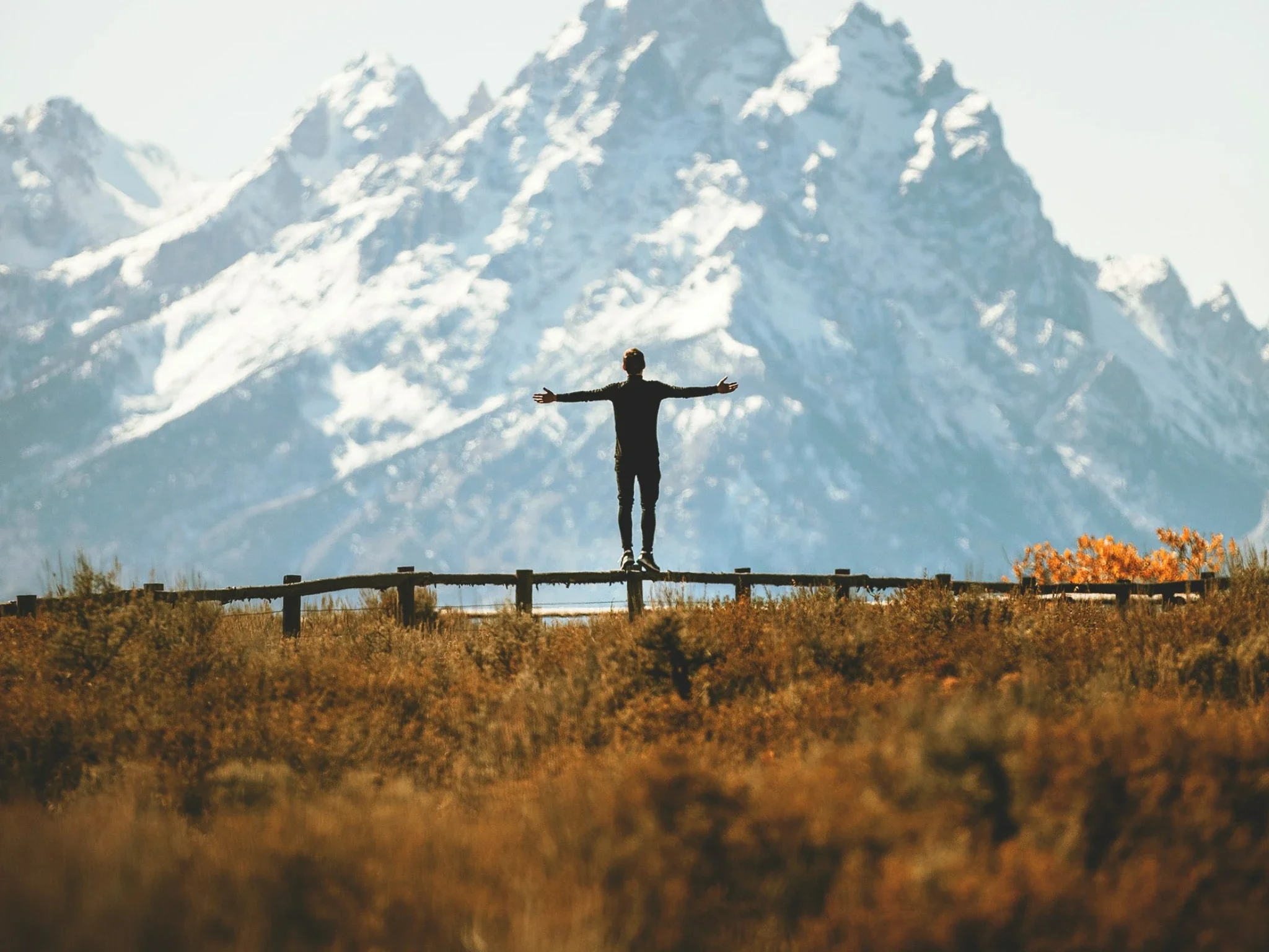 A person standing on a small bridge with arms outstretched, facing a mountain range covered in snow.