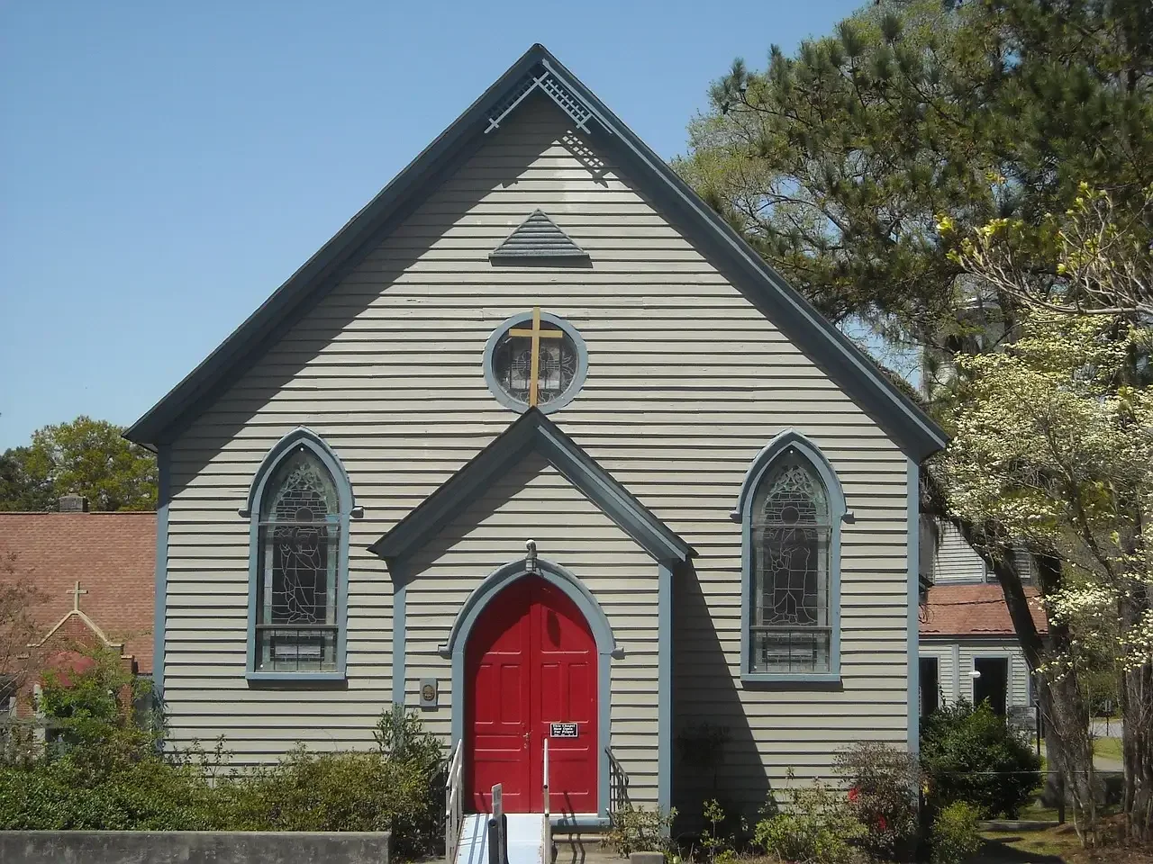 A small church with a steep gabled roof and beige siding, featuring arched stained glass windows and a prominent red front door, surrounded by trees and a clear blue sky.