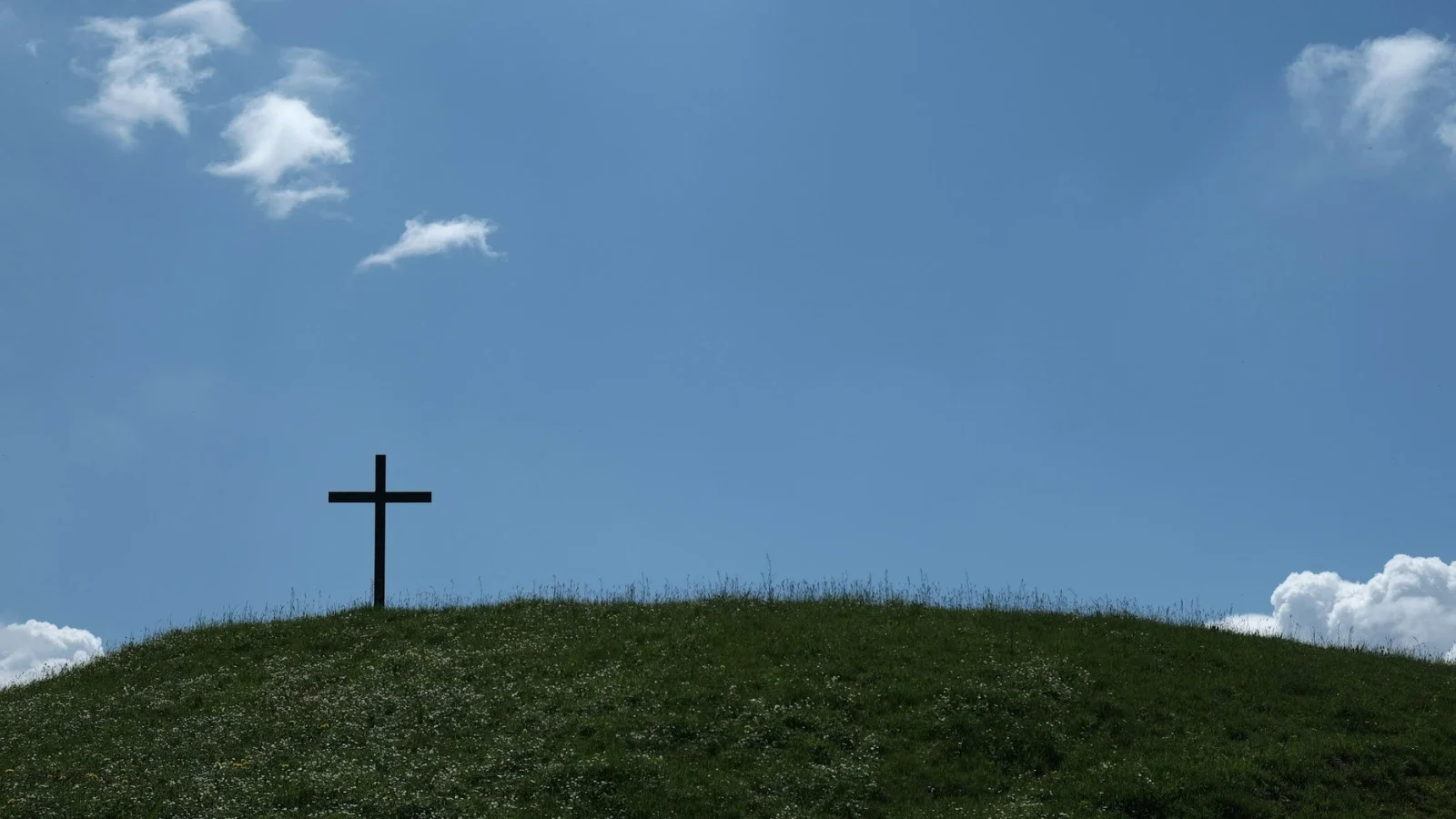 A black cross on a grassy hilltop with a blue sky and white clouds in the background.