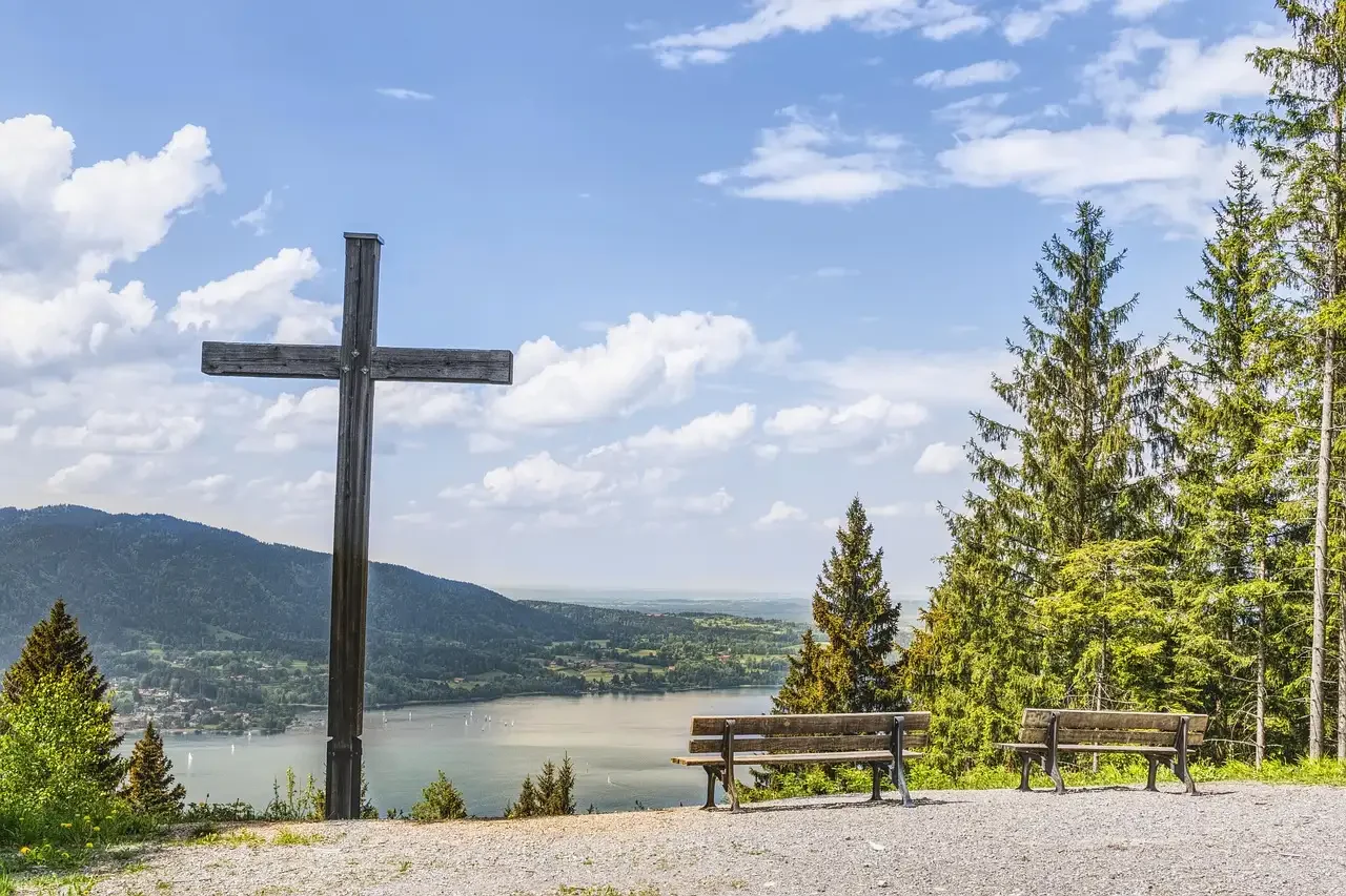 A wooden cross on a hill overlooking a river with sailboats, surrounded by tall evergreen trees and a mountain in the background under a partly cloudy sky.