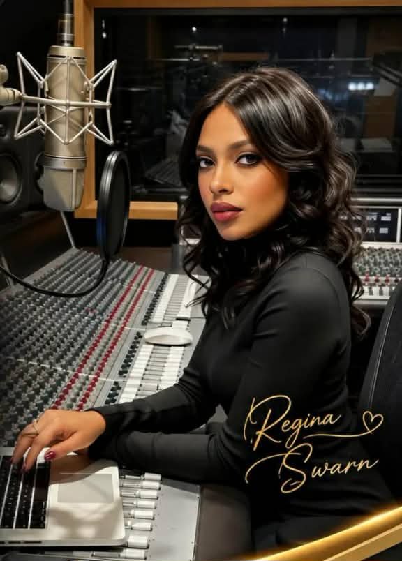 A woman with long dark curly hair in a recording studio, sitting at a sound mixing console with a microphone in front of her, wearing a black top.