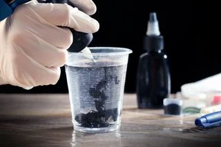 A gloved hand holding a test tube pouring liquid into a clear plastic cup with black particles inside, with various laboratory tools and bottles in the background.