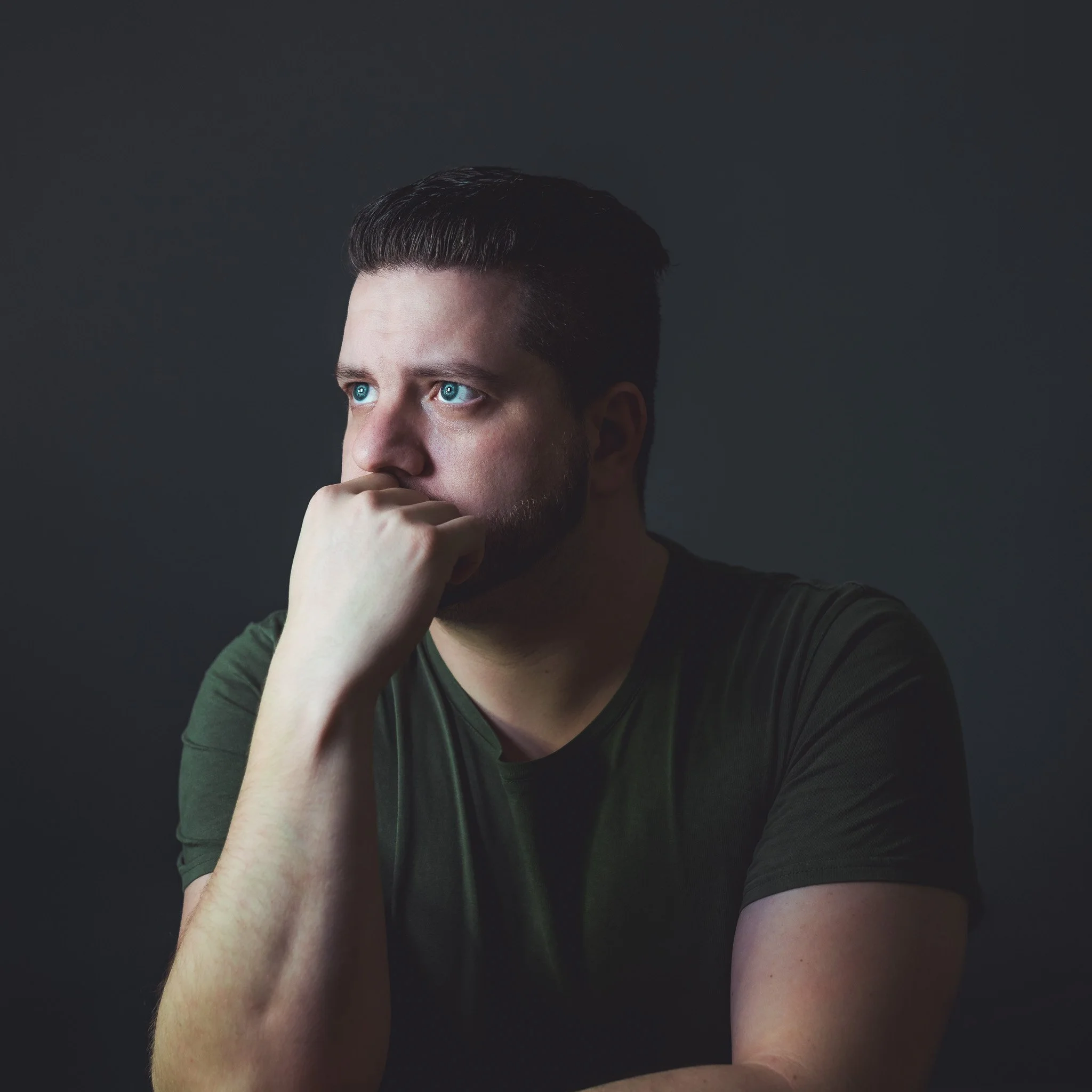 A young man with short dark hair and blue eyes, wearing a dark shirt, is resting his chin on his hand against a dark background.