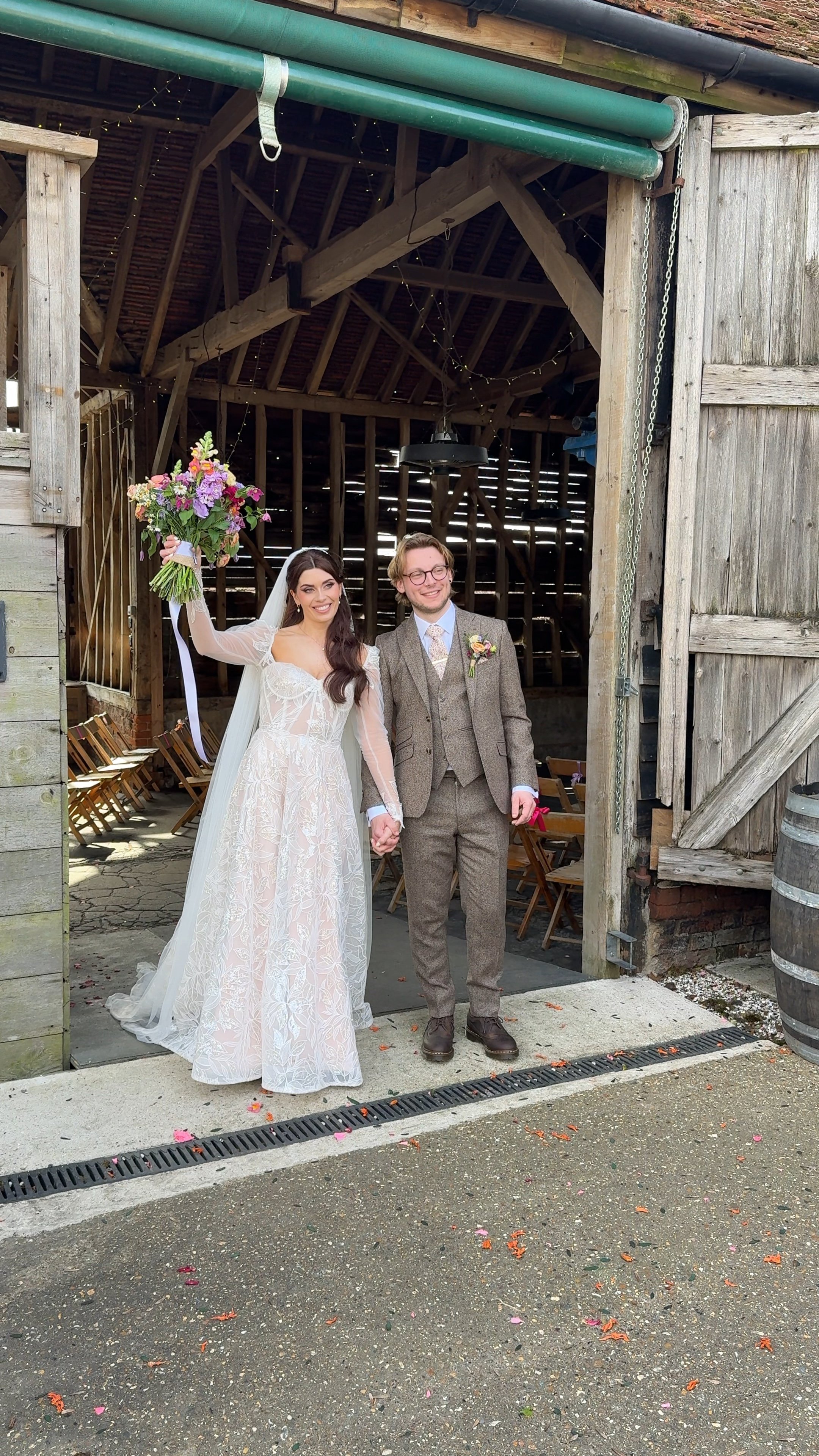 A newly married couple standing hand in hand outside a rustic barn, celebrating their wedding. The bride is holding a bouquet of flowers and smiling, while the groom is in a suit with glasses and also smiling. The barn has wooden doors and seating inside, and there are string lights hanging from the ceiling.