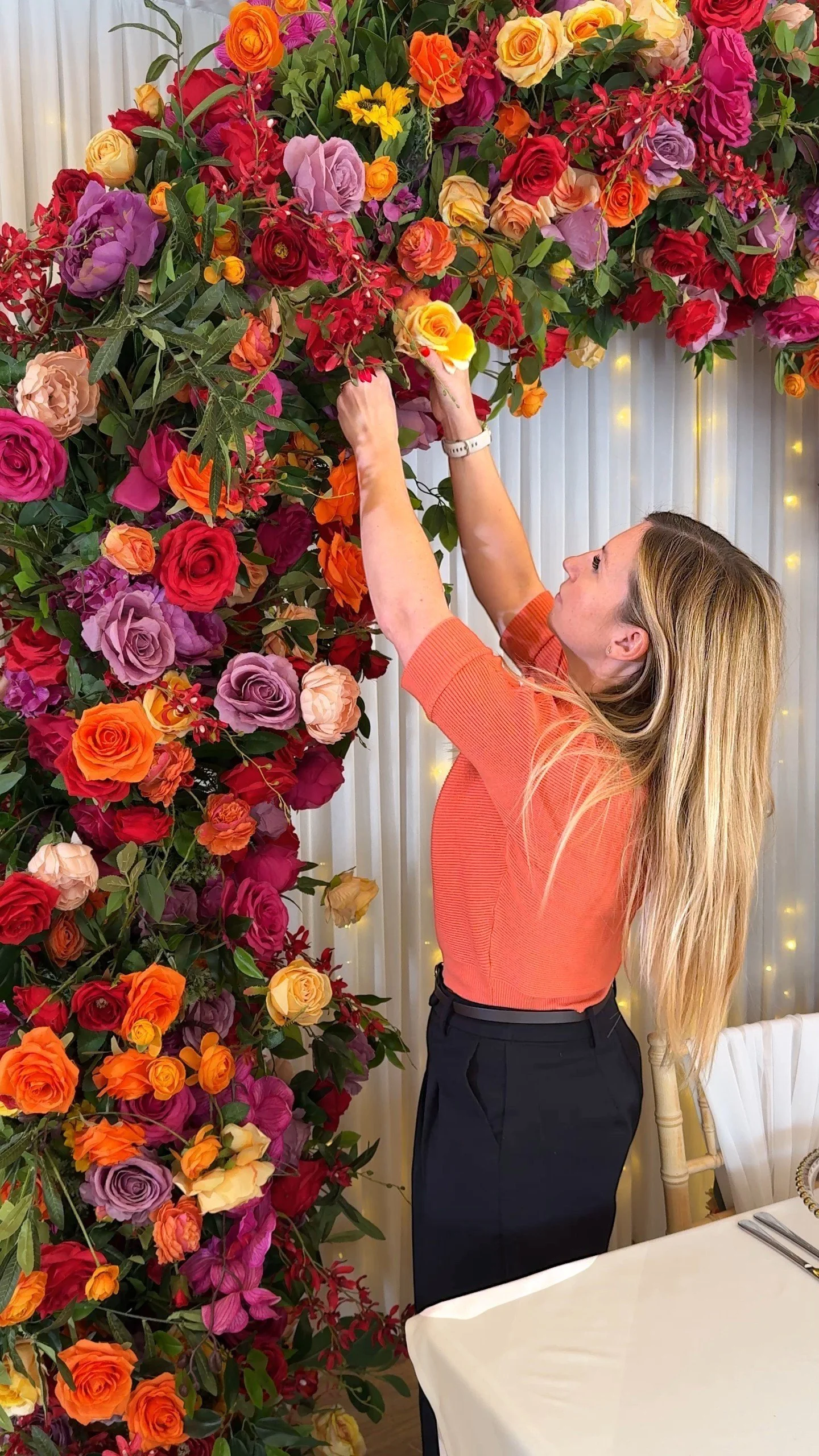 A woman arranging a vibrant, large wall of colorful roses and flowers at an indoor event or celebration.
