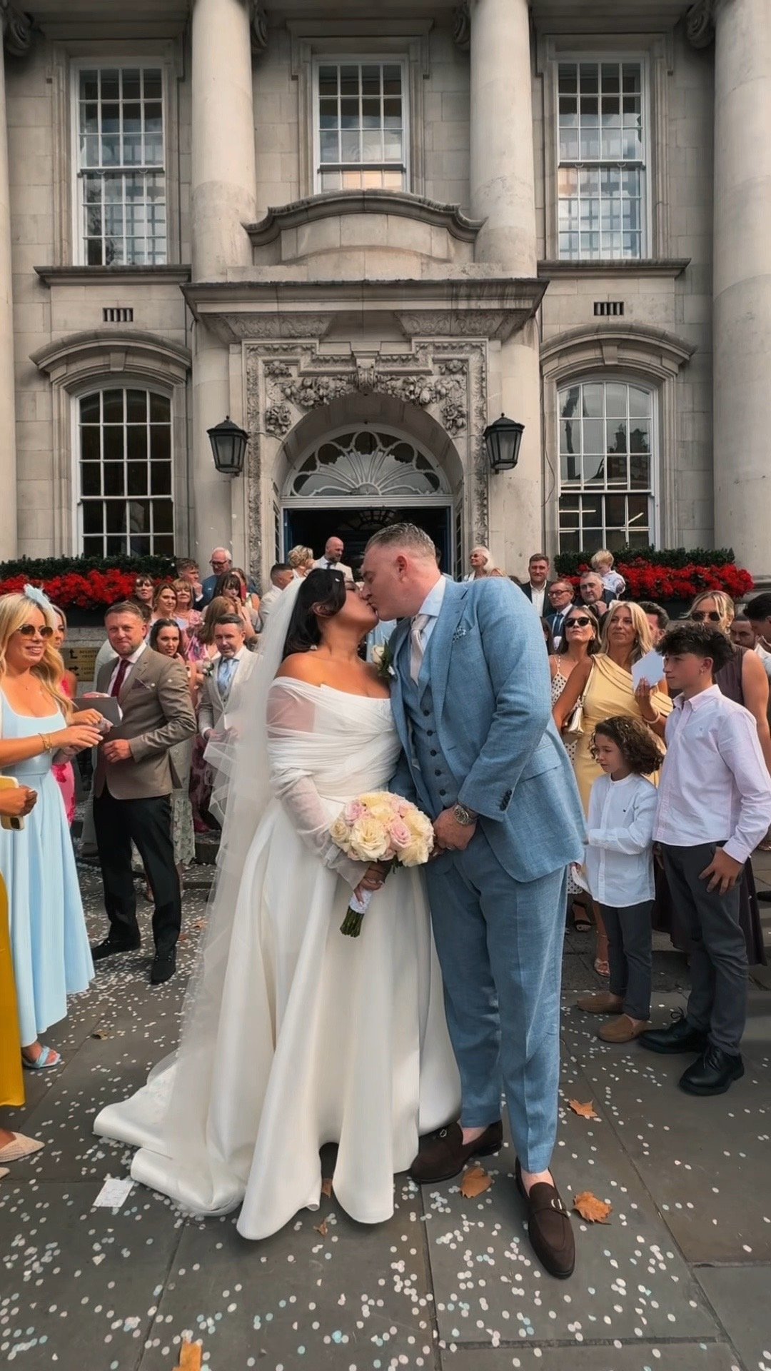 A bride in a white wedding dress and a groom in a blue suit kiss in front of a crowd outside a grand building with columns, with people dressed in formal attire around them.