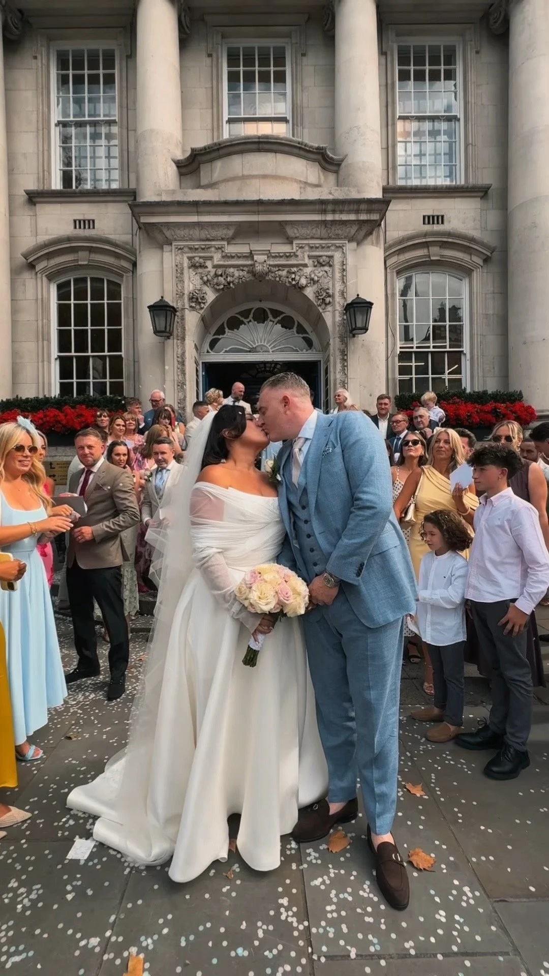 A bride in a white wedding dress and a groom in a blue suit kiss in front of a crowd outside a grand building with columns, with people dressed in formal attire around them.