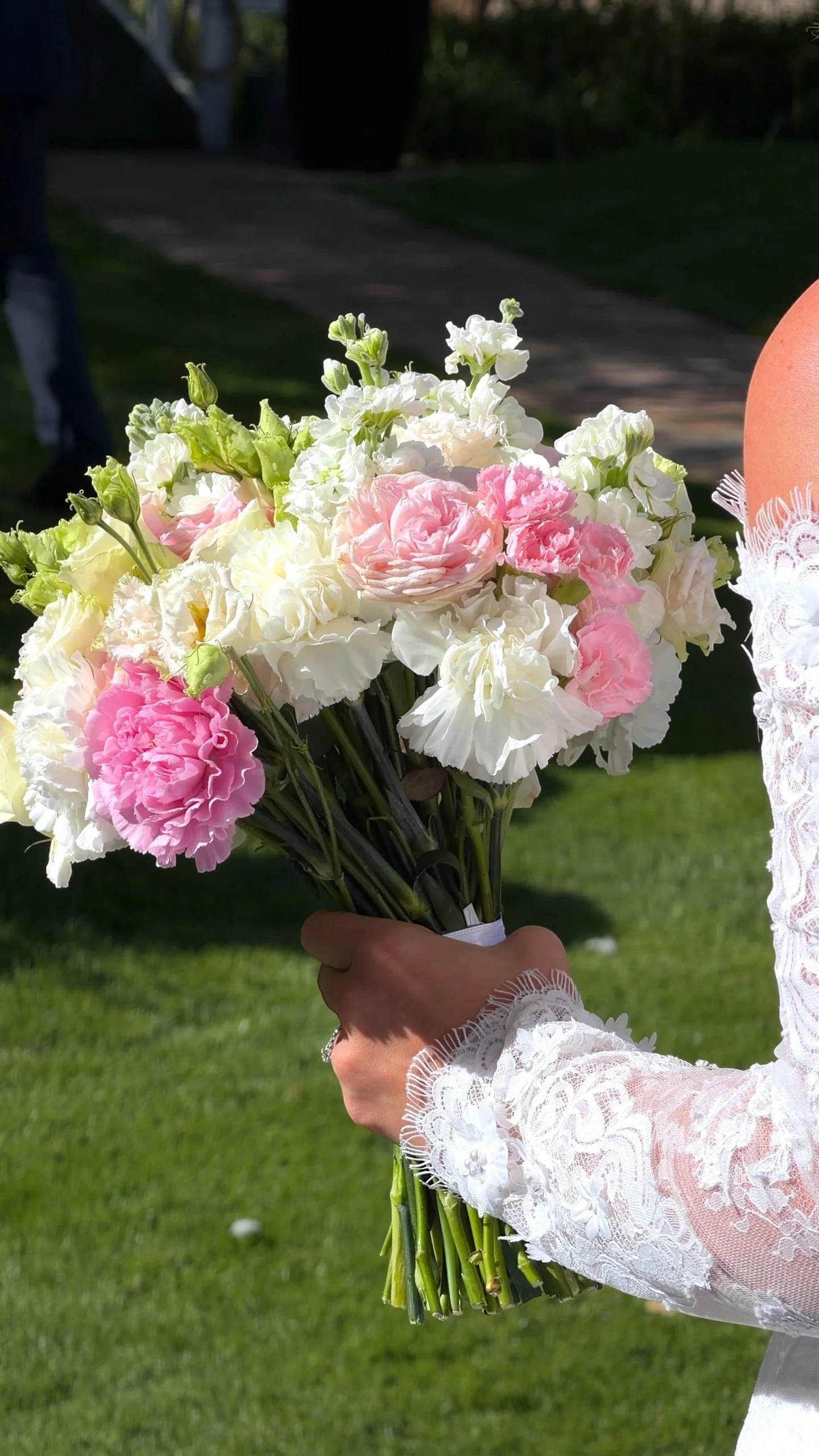 Person holding a bouquet of pink and white flowers outdoors, wearing white lace clothing with a green lawn background.