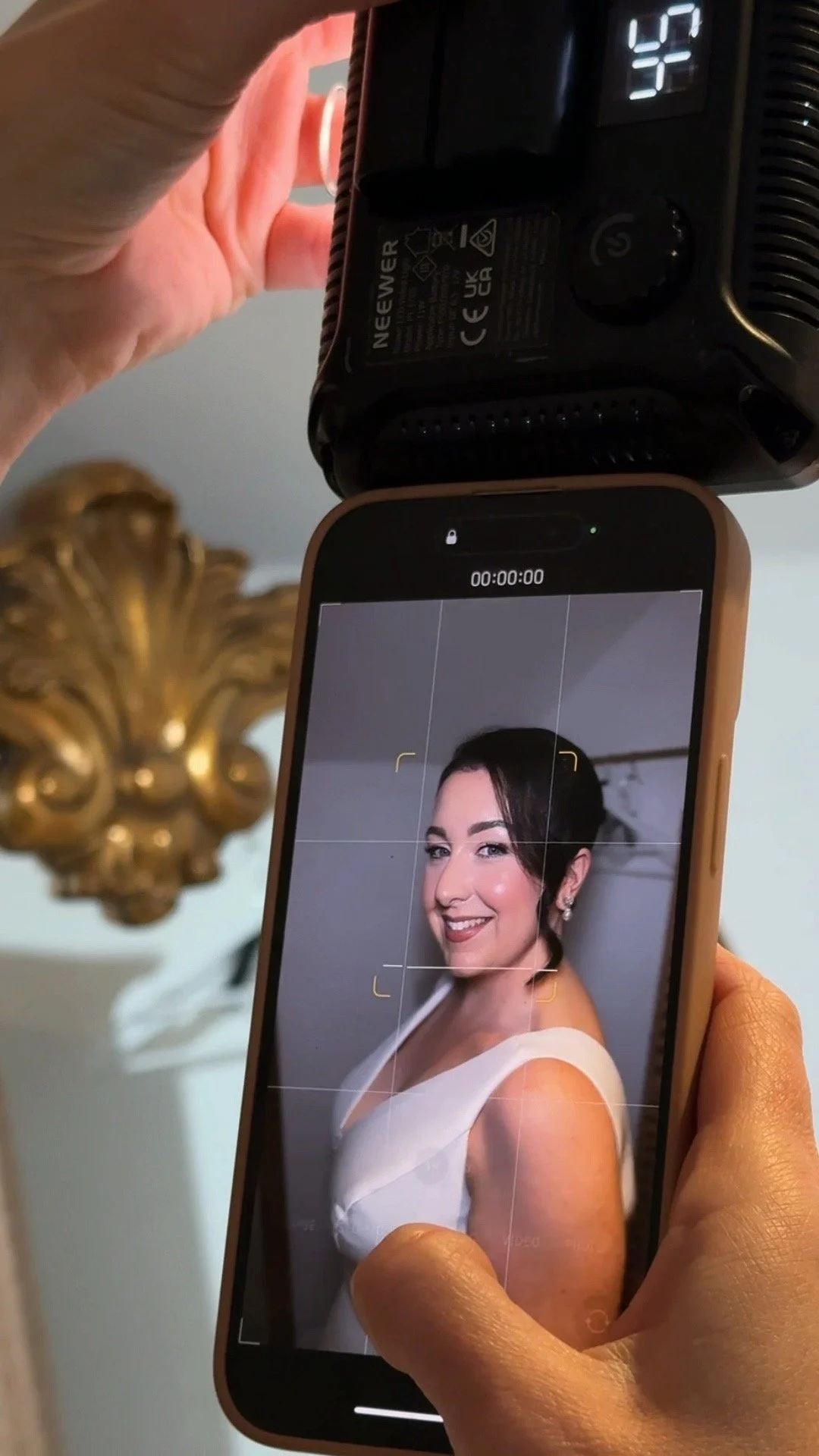 Person taking a photo of a woman in a white dress using a smartphone with a camera app open, and a black electronic device with a digital display in the background.