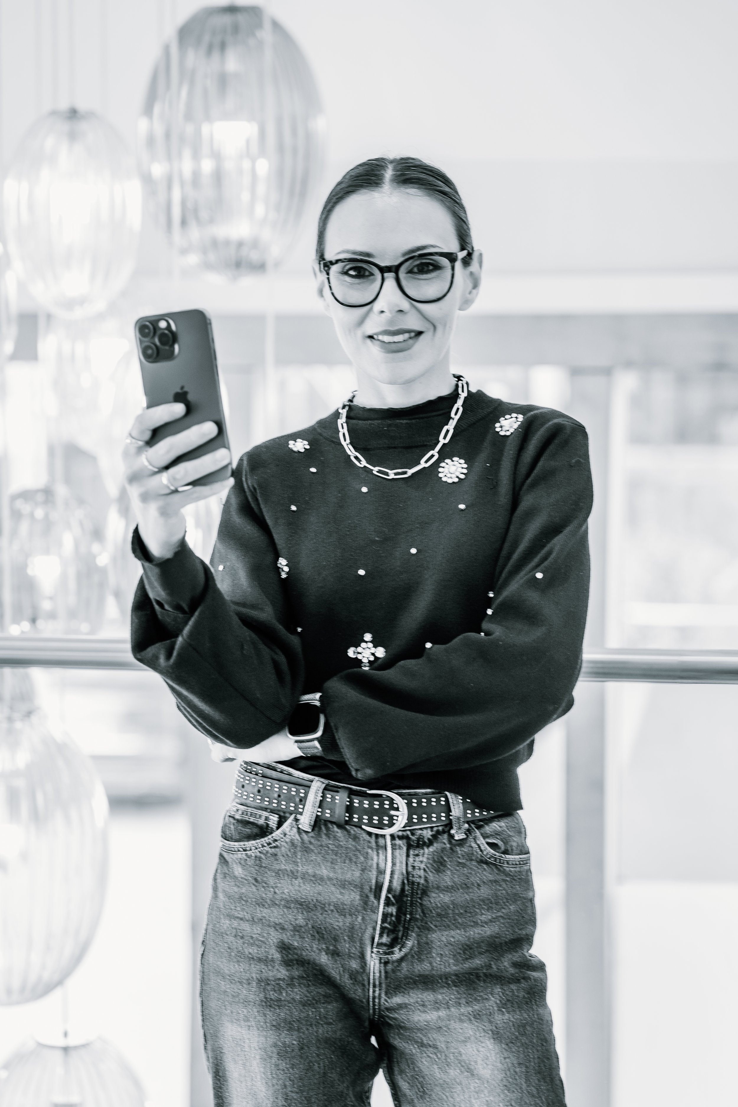 A woman with glasses and a chain necklace takes a selfie, wearing a dark sweater with embellishments and high-waisted jeans, standing indoors in front of large decorative lamps.