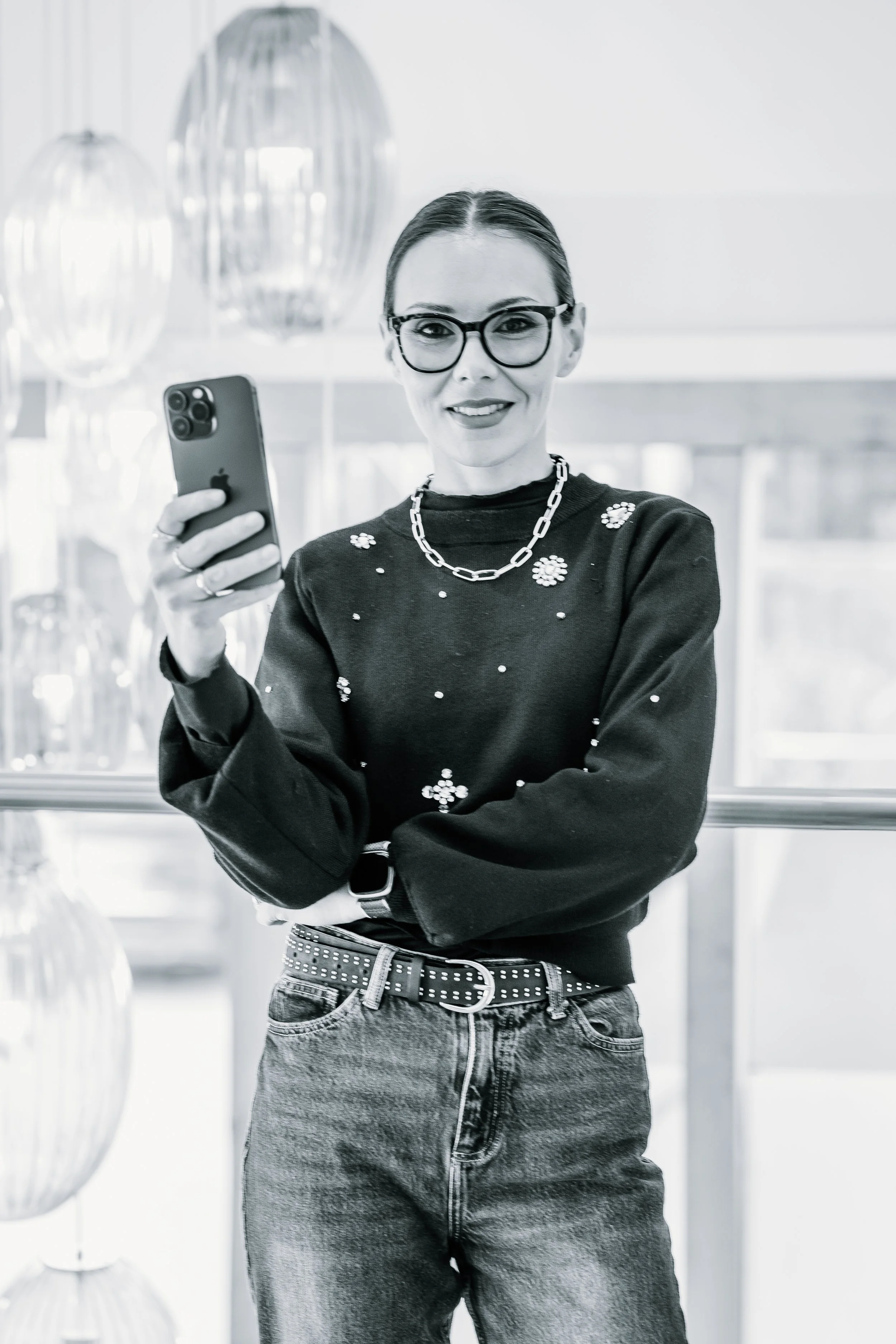 A woman with glasses and a chain necklace takes a selfie, wearing a dark sweater with embellishments and high-waisted jeans, standing indoors in front of large decorative lamps.